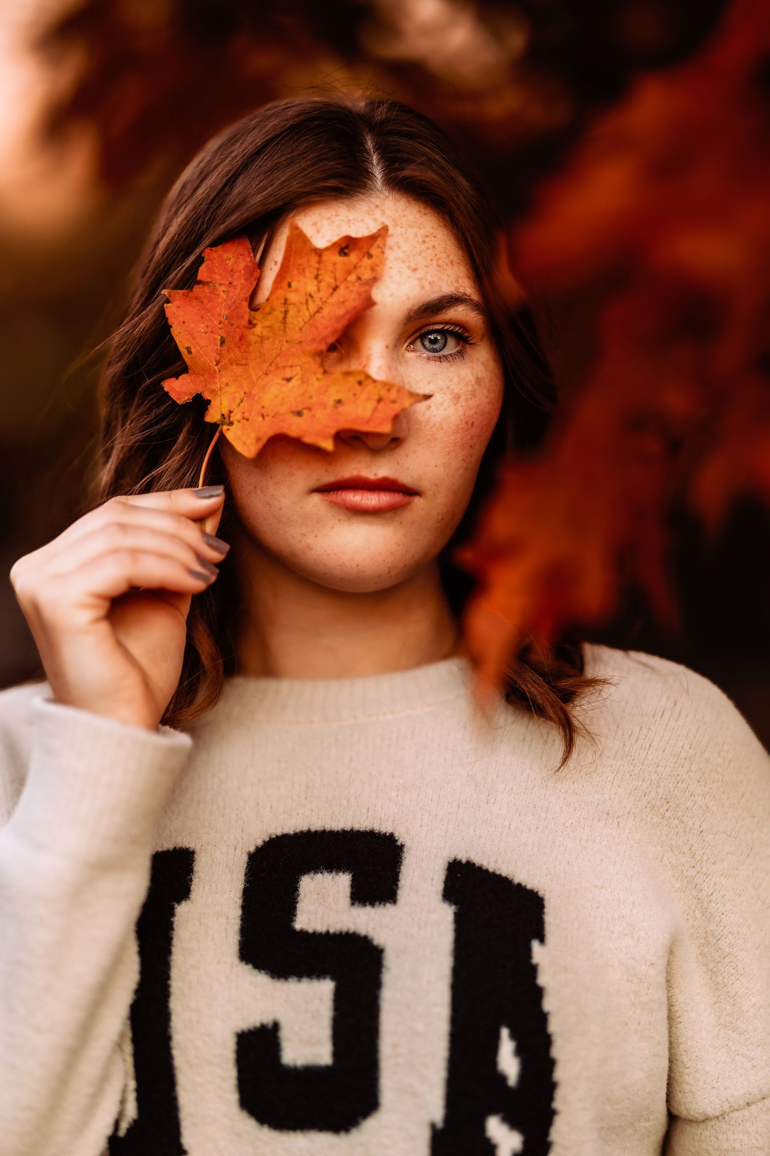 A fall portrait with an orange maple leaf held up against autumn foliage background and a cream-colored USA sweater.