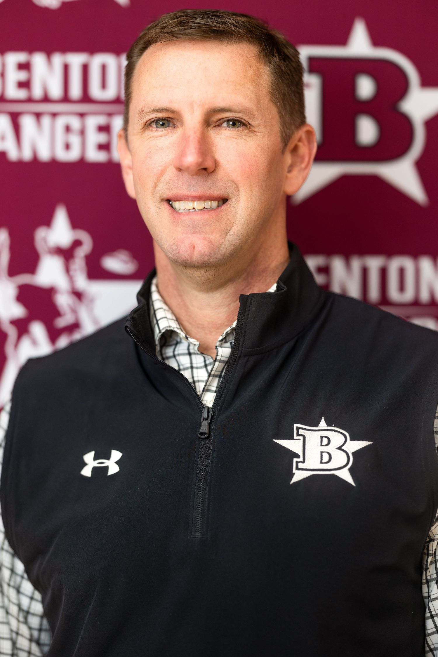 Athletic coach in Under Armour quarter-zip jacket smiles in front of team logo backdrop.