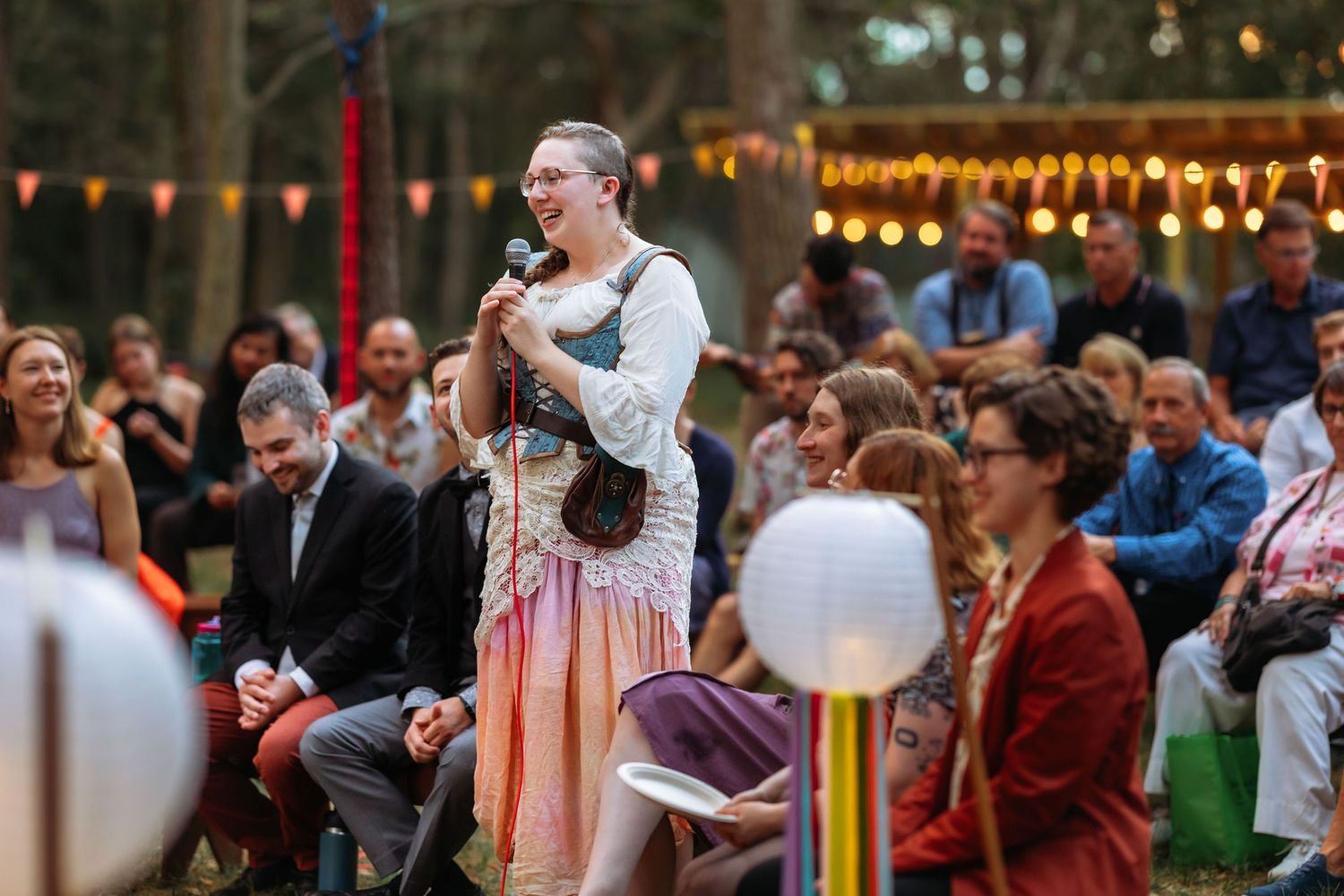 A guest gives a speech at an outdoor wedding reception with string lights and a crowd in the background.