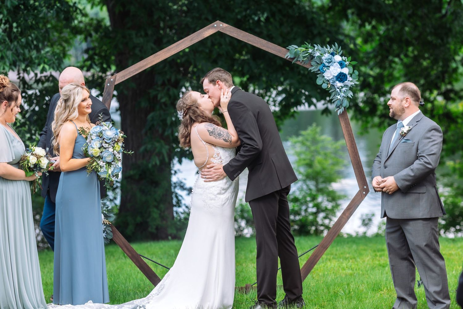 Wedding couple shares first kiss under wooden arch during outdoor ceremony.