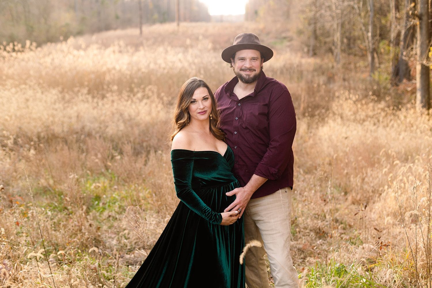 Couple poses together in autumn field, woman in green velvet dress and man in burgundy shirt with hat.