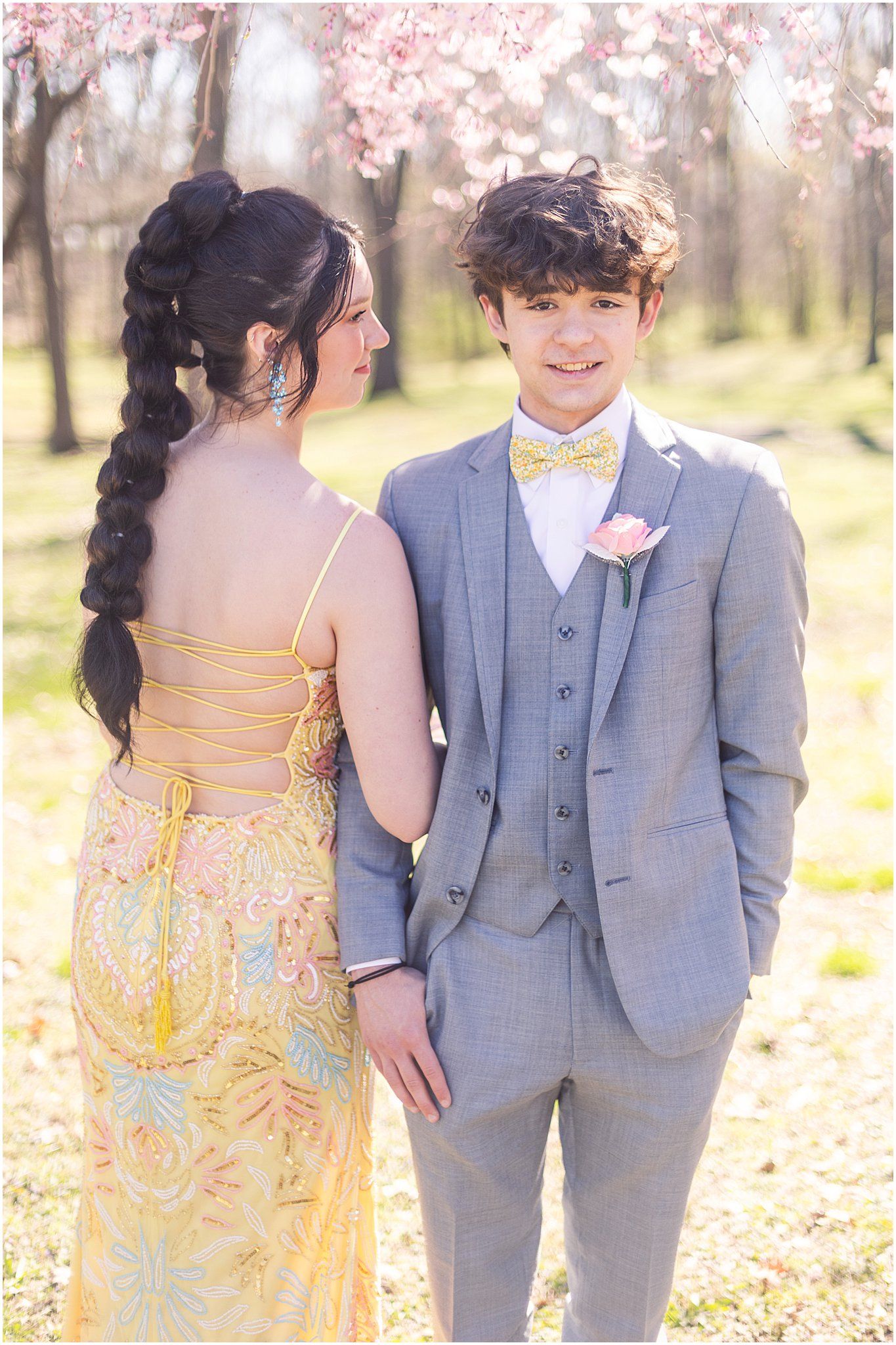 Couple in formal attire standing outdoors with cherry blossoms, wearing yellow and grey.