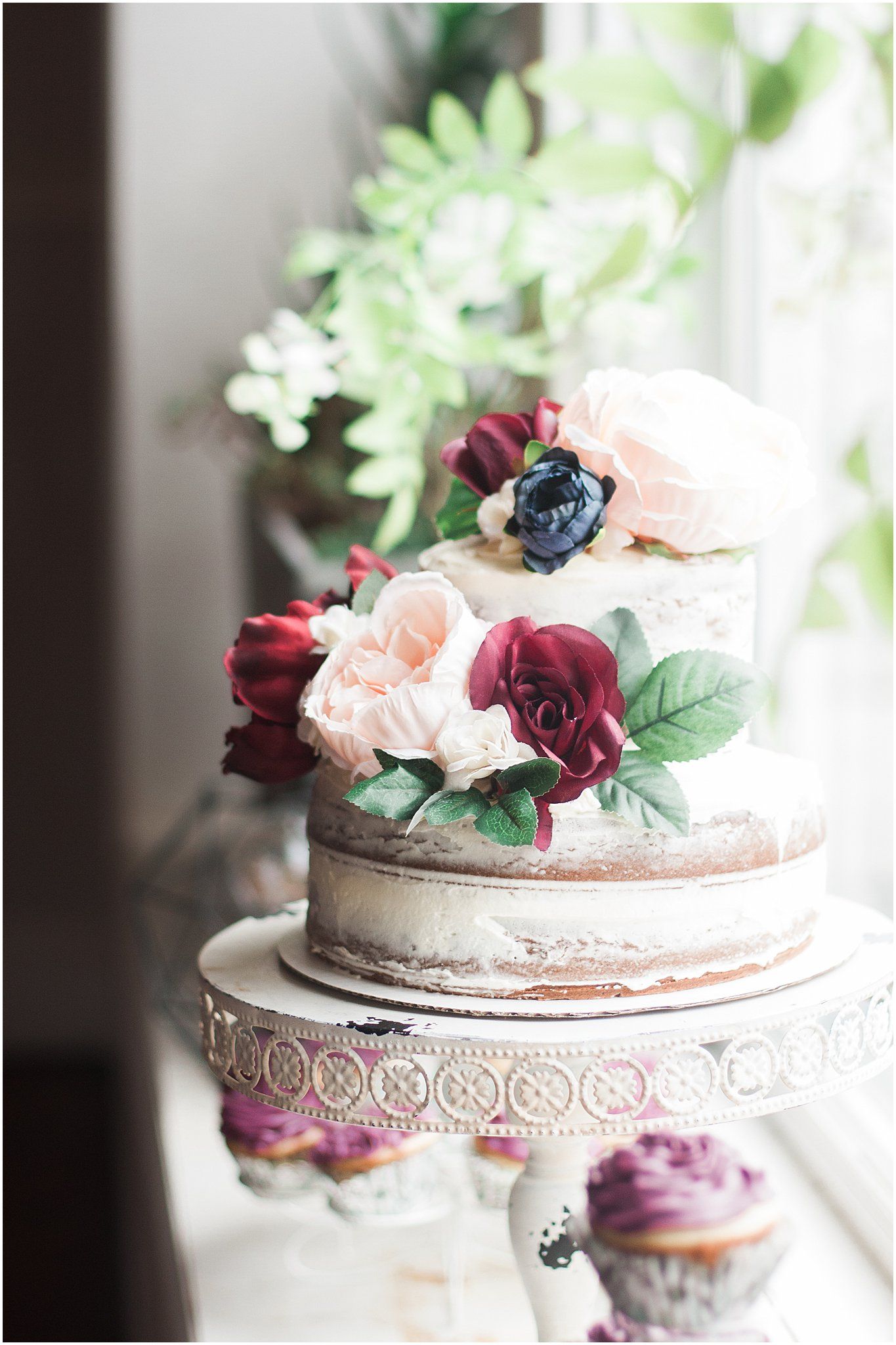 A rustic naked wedding cake decorated with burgundy and blush roses on an ornate white cake stand.