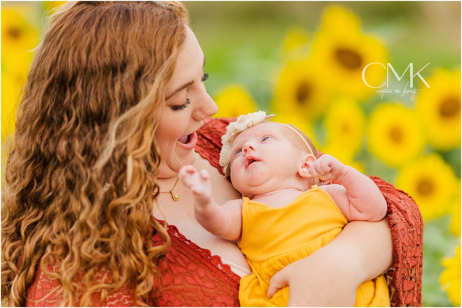 Close-up portrait of parent cradling baby wearing yellow against backdrop of bright sunflowers.