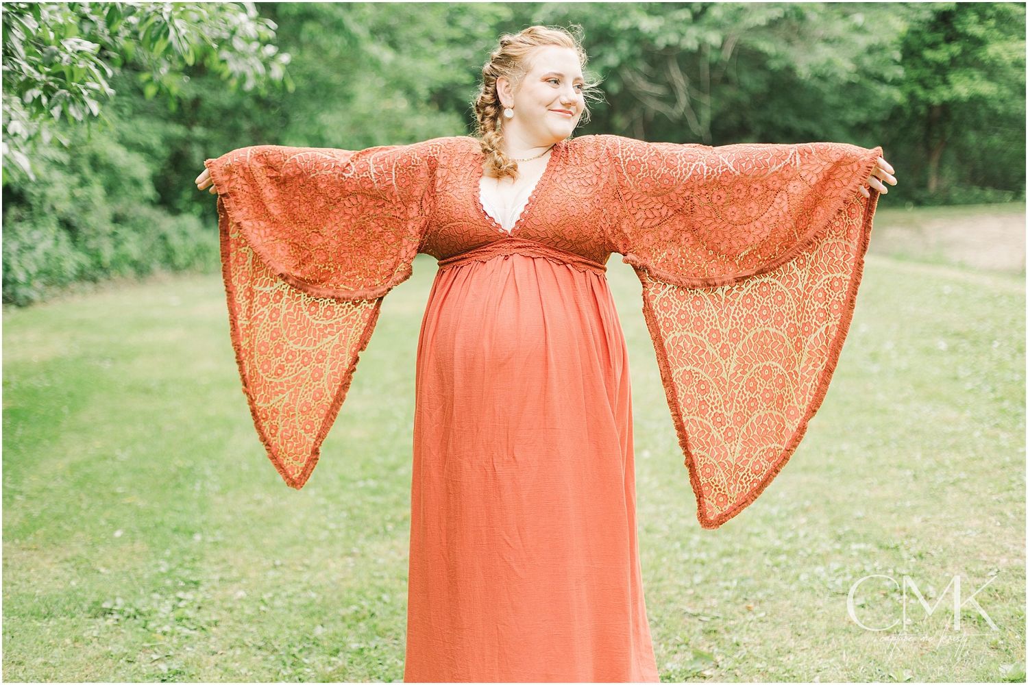 Maternity portrait showcasing dramatic bell sleeves of coral lace dress in garden setting.