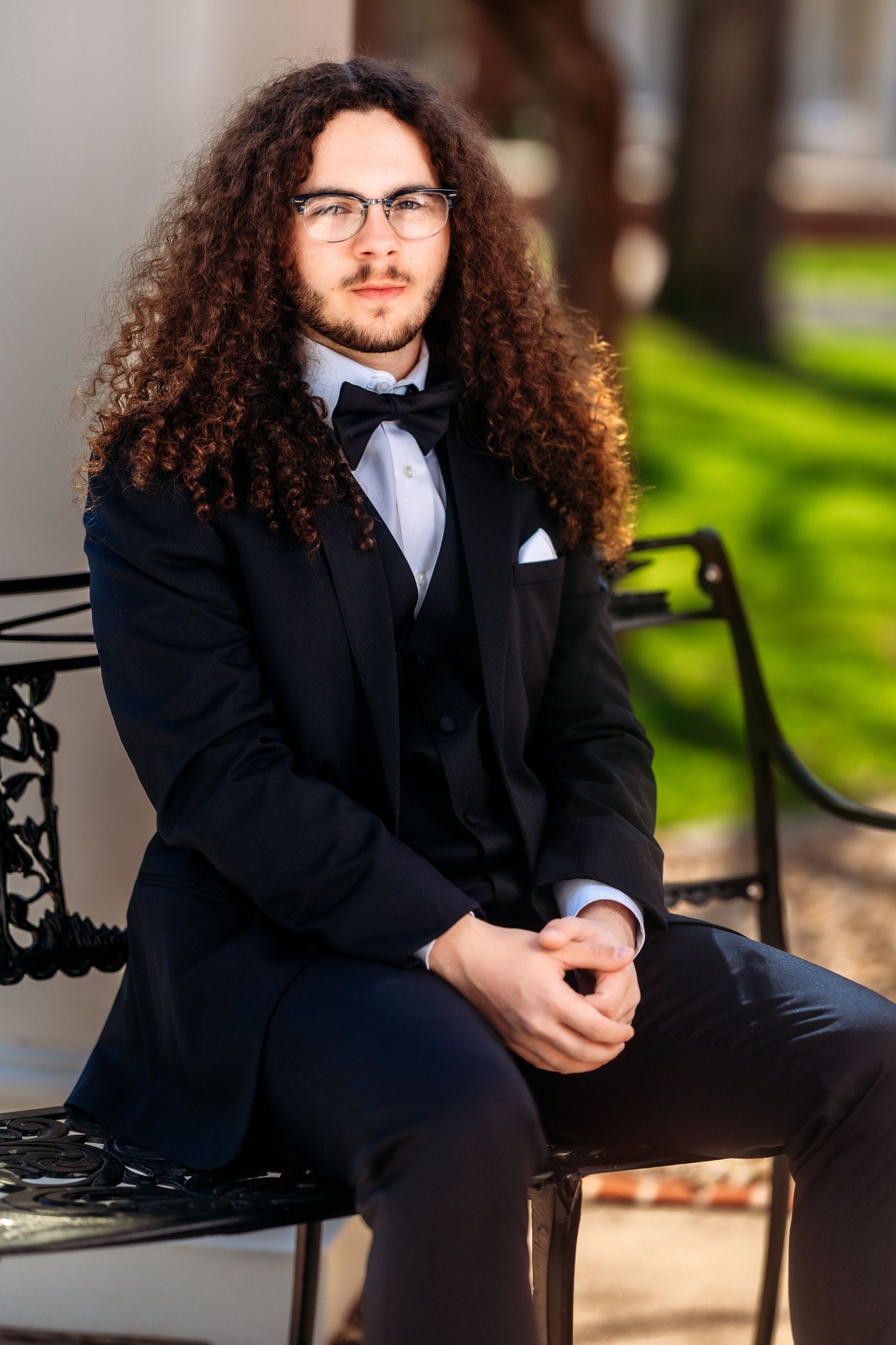 Professional portrait of someone in a black suit and white shirt sitting on a decorative bench outdoors.