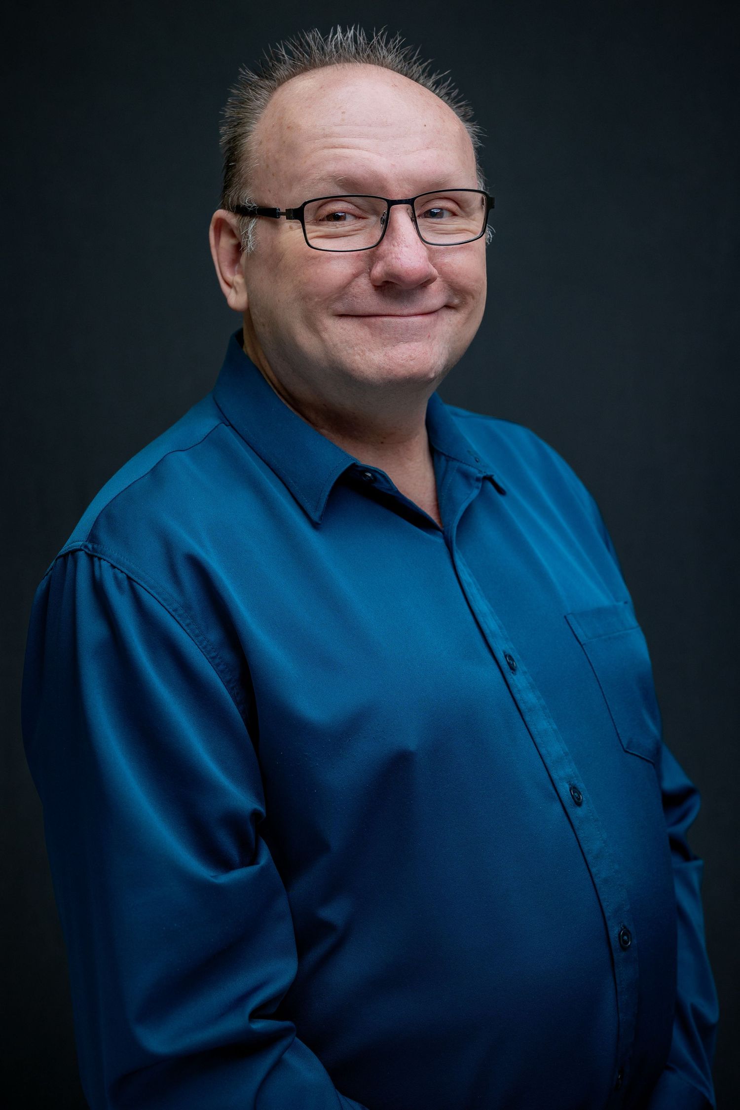 Business portrait of man in blue button-down shirt and glasses against dark backdrop.