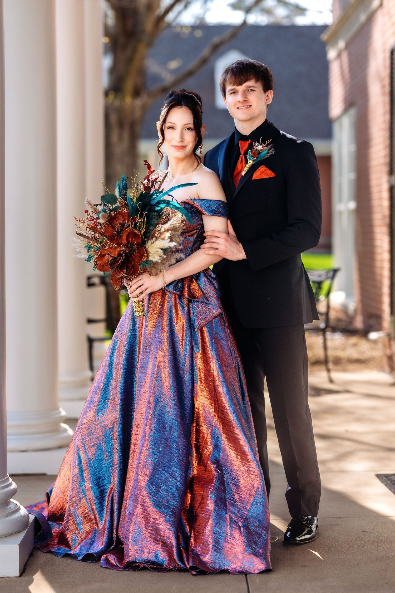 Formal couple poses in black tuxedo and iridescent blue gown with autumn-themed bouquet.