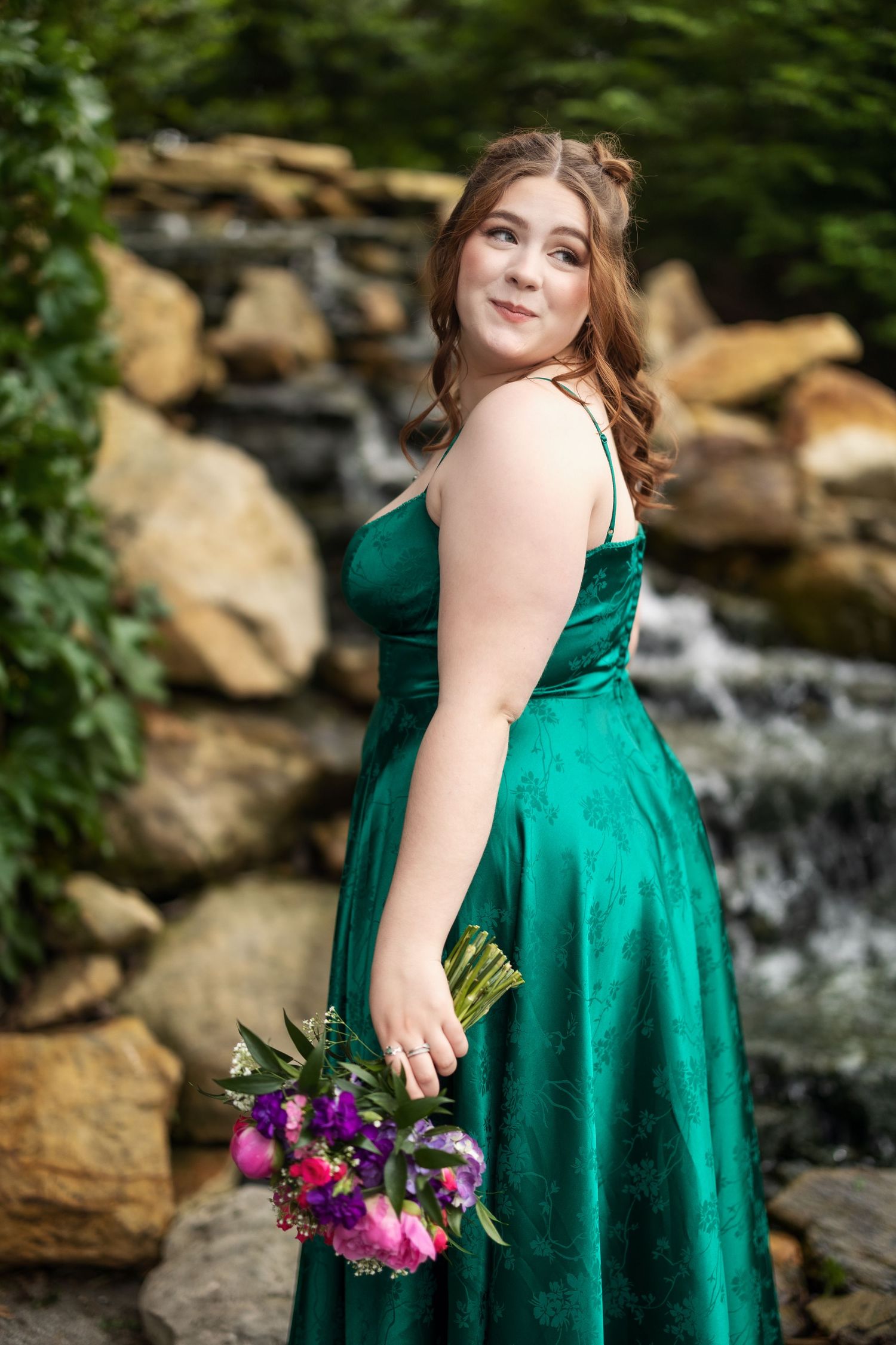Young woman in emerald green satin prom dress holding pink and purple bouquet, posing by rocky waterfall outdoors.