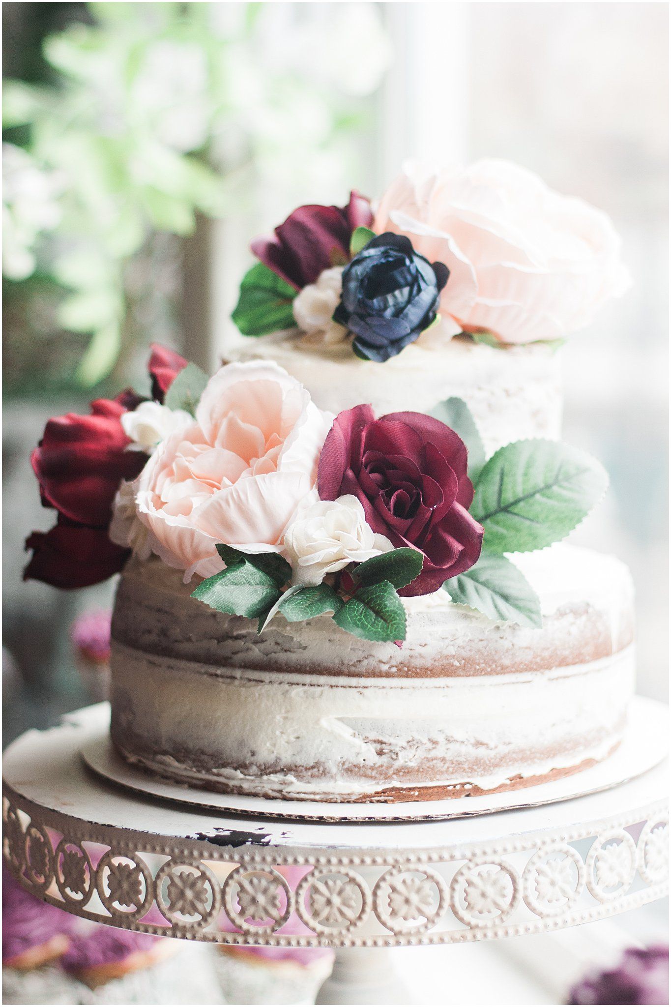 A rustic semi-naked cake adorned with burgundy and blush roses on a decorative stand.