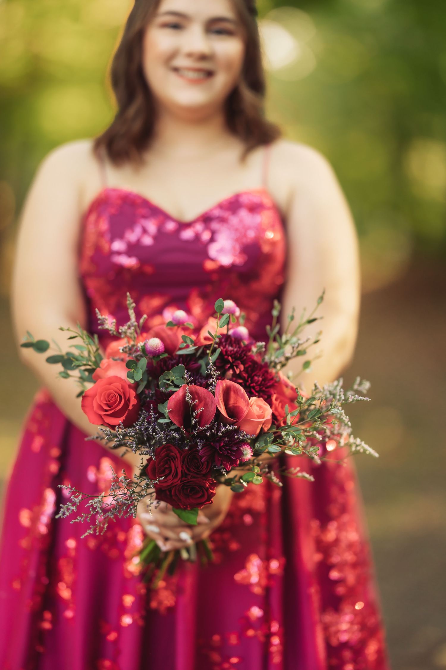 A person in a sparkly red formal dress holds a vibrant bouquet of red roses and greenery against a blurred outdoor background.