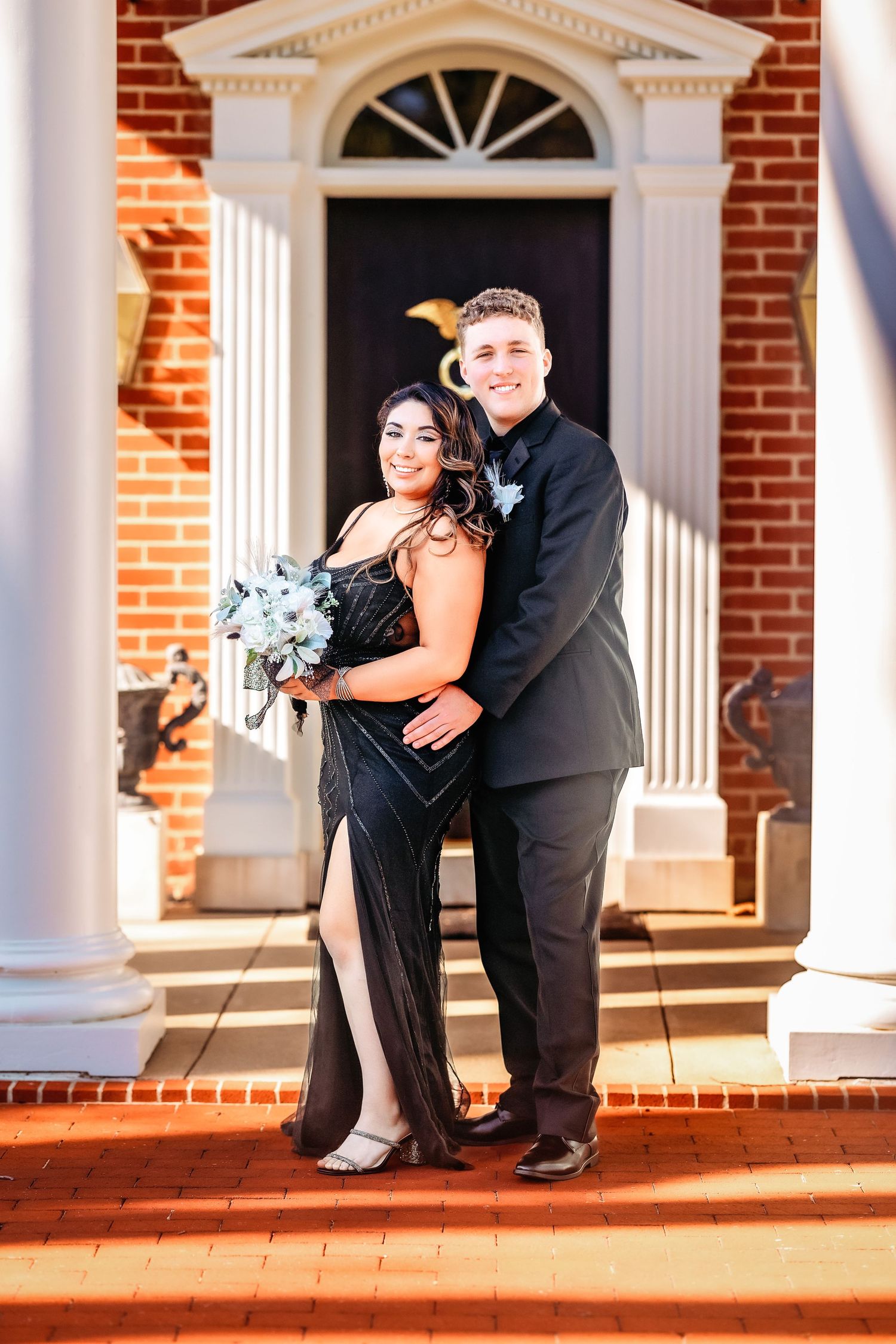 A couple in formal attire shares a romantic moment on a brick porch with white columns and doorways.