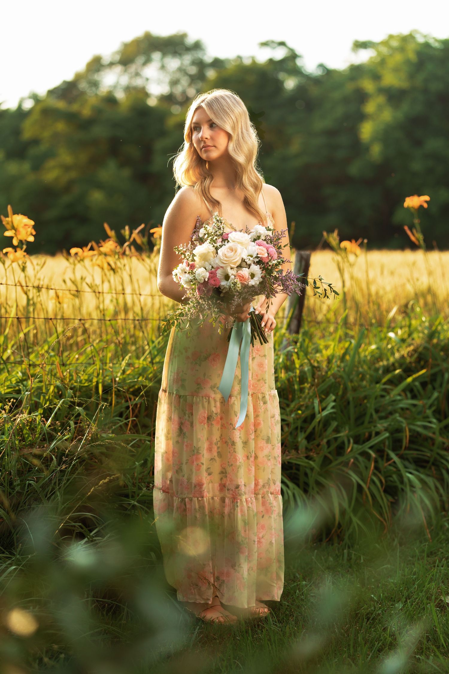 Blonde woman in floral maxi dress holding bouquet in golden hour field.