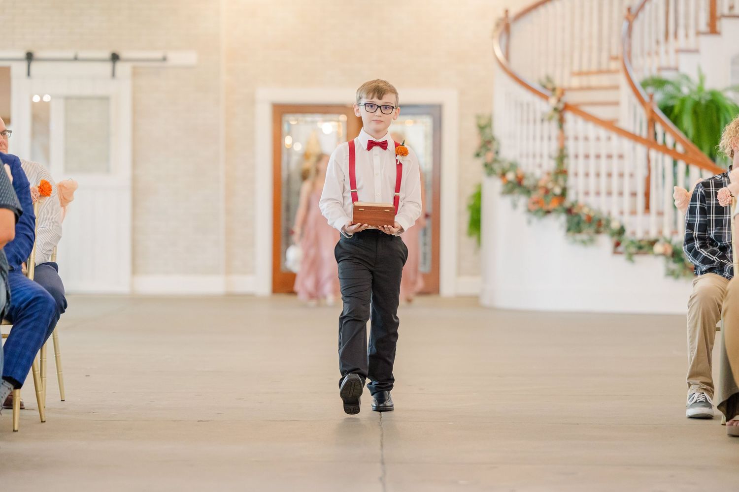 Young ring bearer in suspenders and bow tie walks down aisle during wedding ceremony.