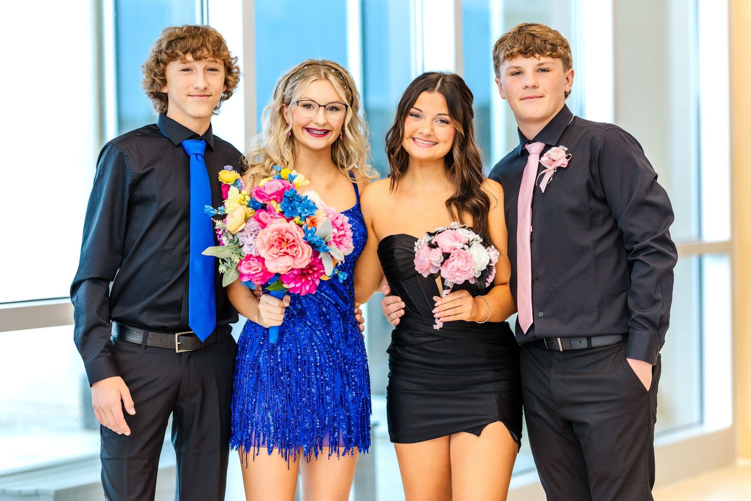 Group photo of four people in formal attire holding colorful bouquets in a bright hallway.