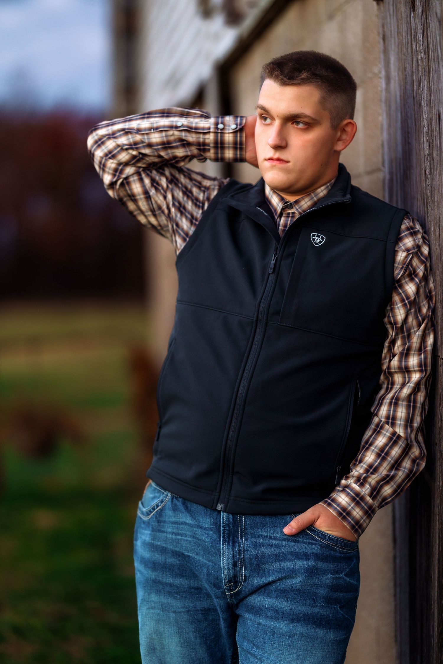 Person wearing a black vest and plaid shirt leaning against rustic wood background.