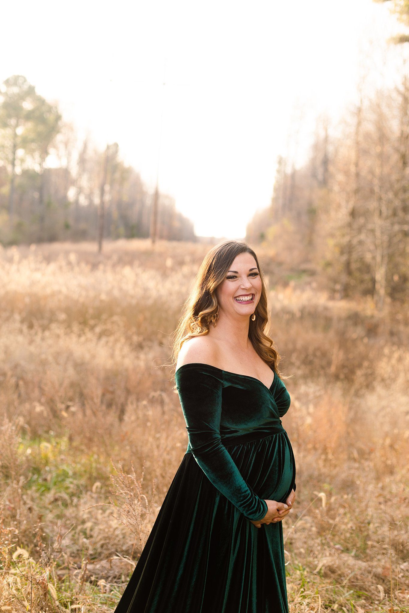 Joyful maternity portrait in emerald velvet gown against warm sunset lighting.