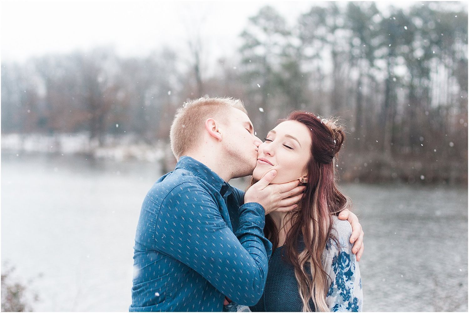A romantic winter photoshoot by a lake features a couple in coordinating blue outfits sharing a tender moment.