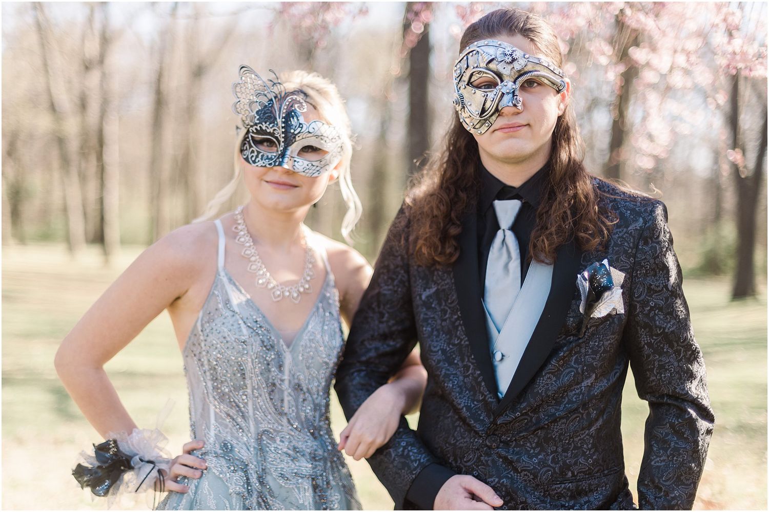 Masquerade-themed formal portrait with couple in blue gown and dark suit under spring blossoms.