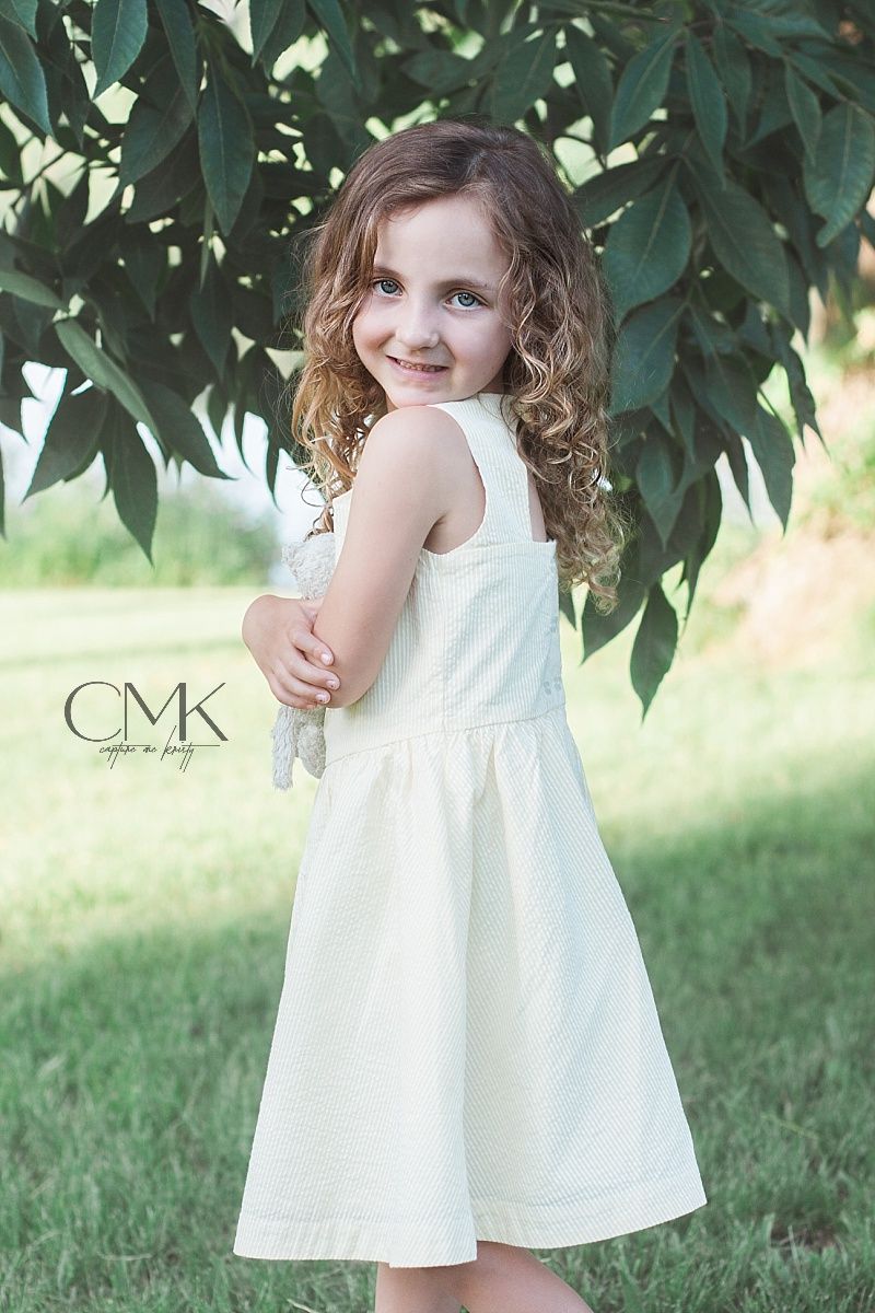 Sweet portrait of child in white summer dress posing against green garden backdrop.