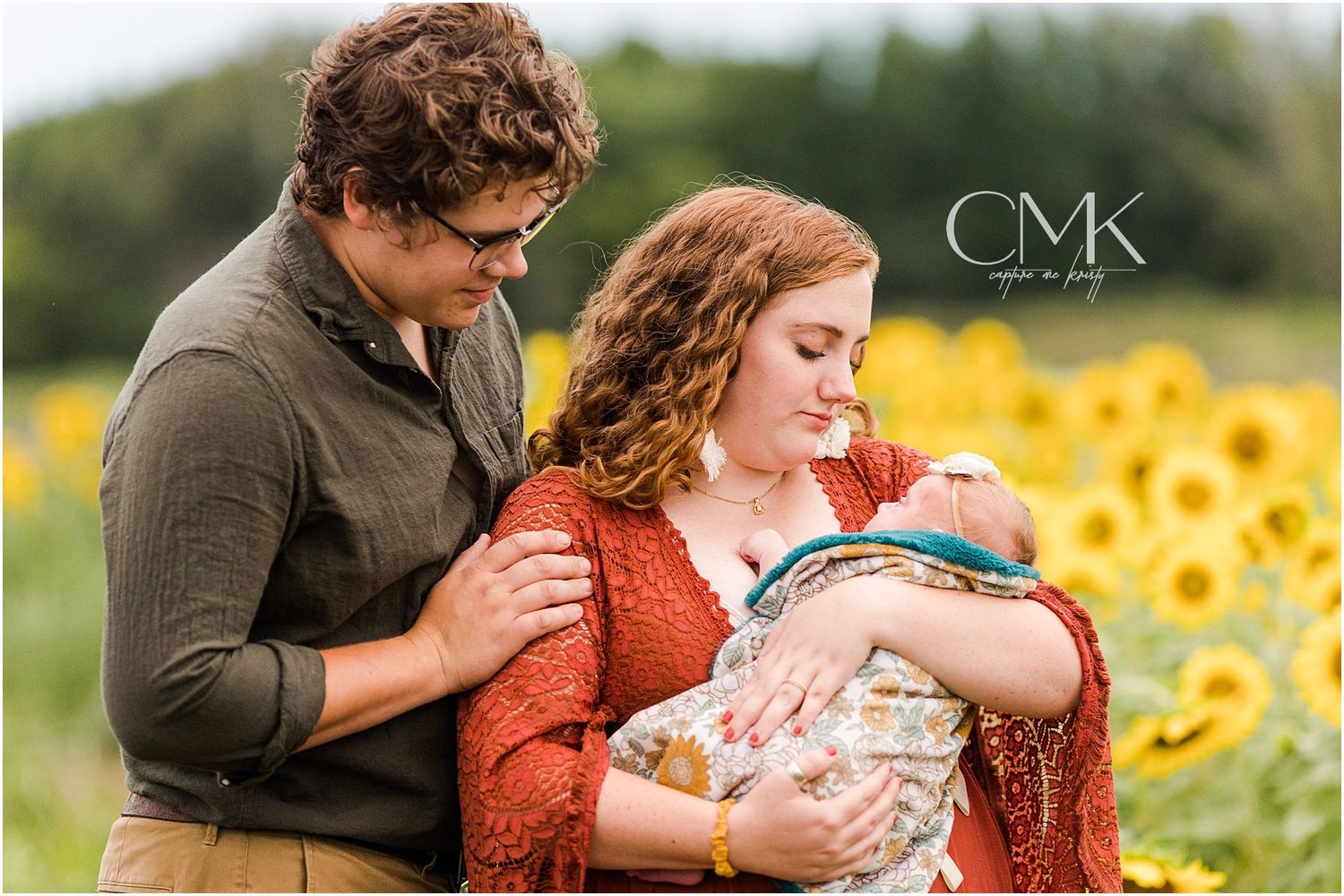 Family portrait in sunflower field with baby wrapped in patterned blanket.