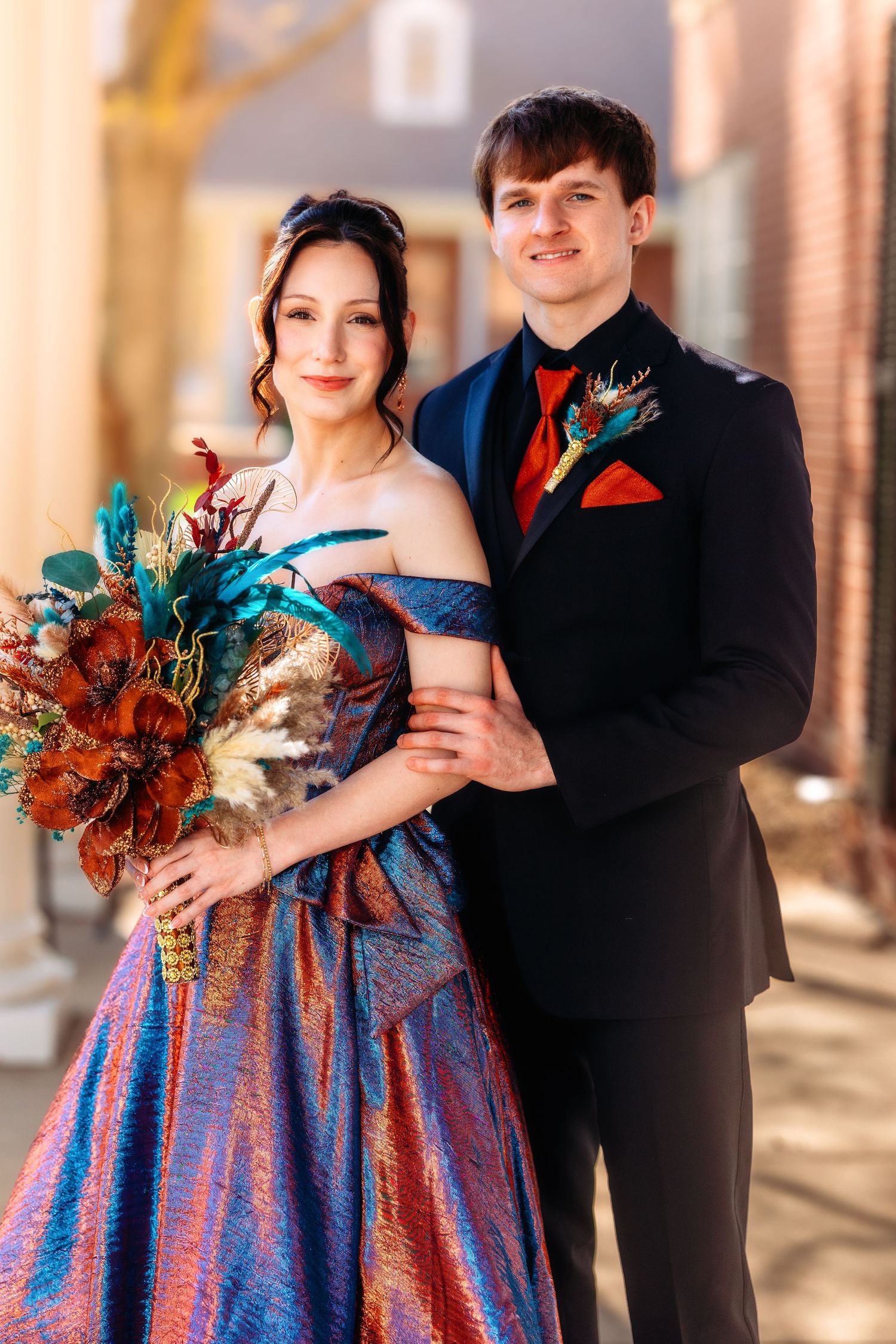 A couple in formal attire poses together, with the woman wearing a blue and pink dress holding an autumn-colored bouquet.
