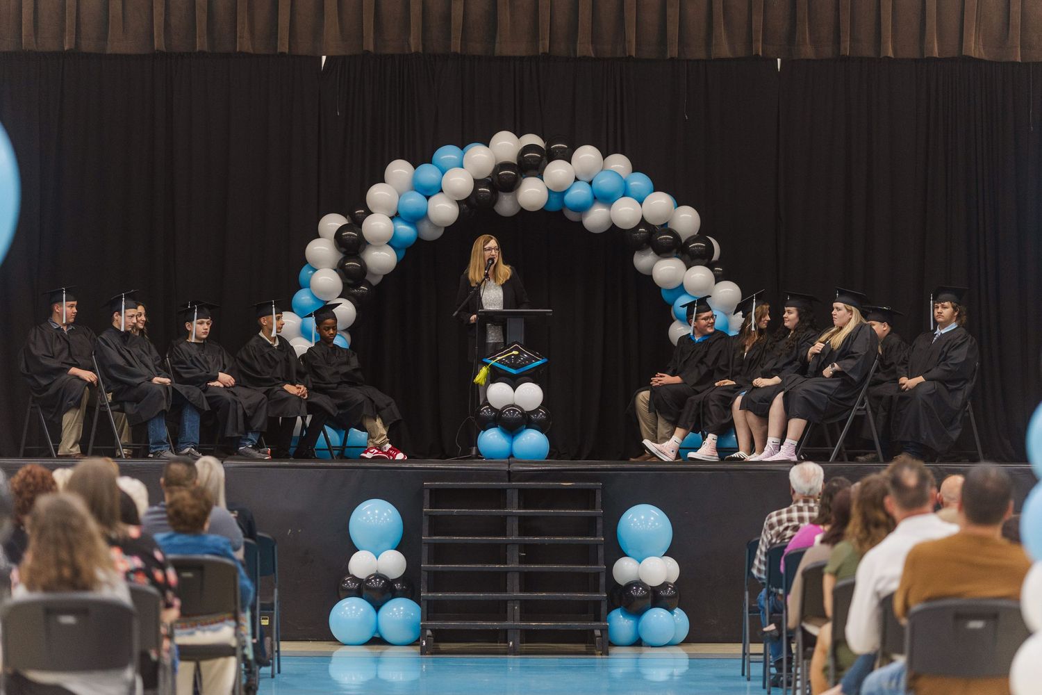 An audience seated in rows watches a performance with blue and white balloon decorations.