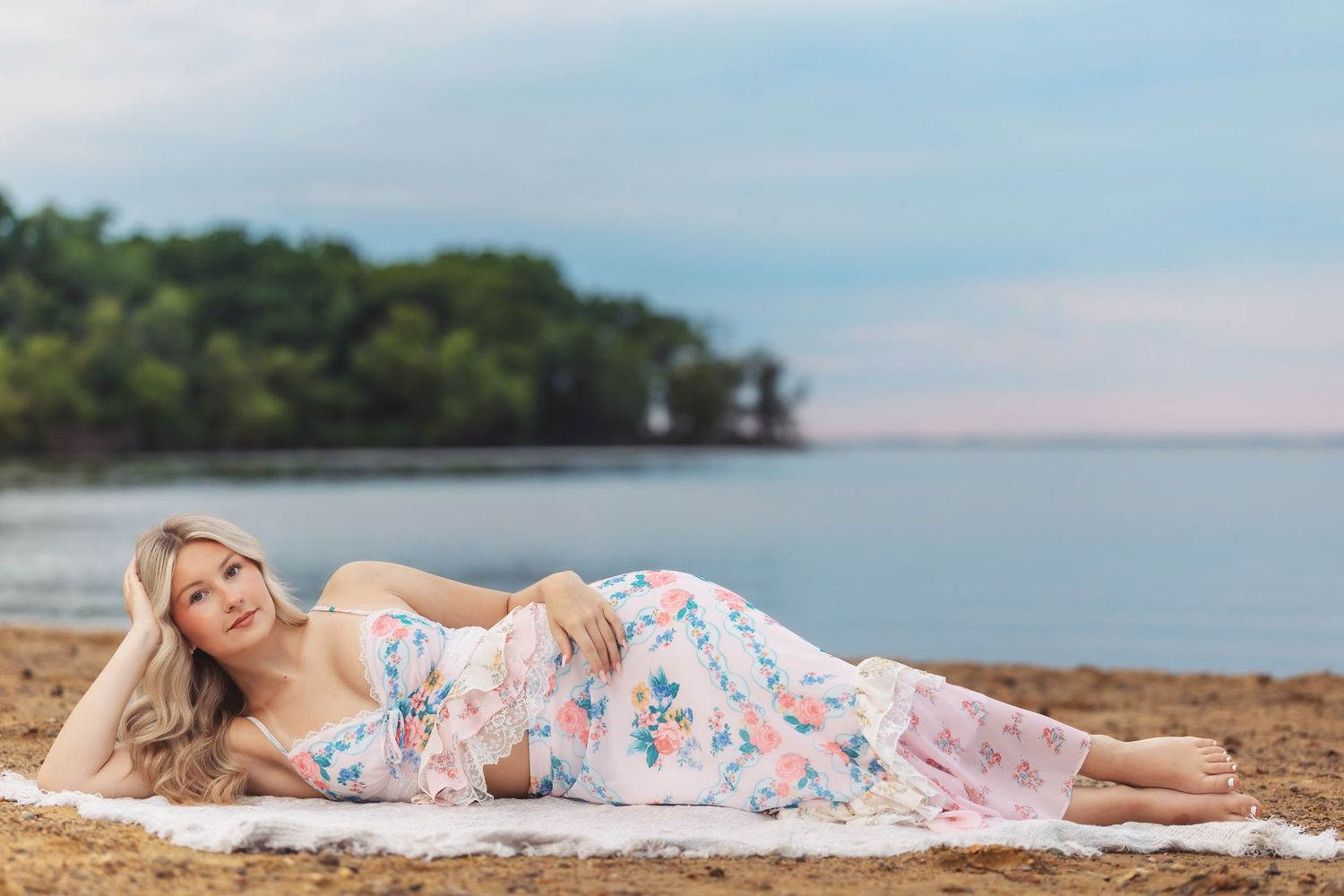 A blonde woman in a floral maxi dress reclines gracefully on a white blanket at a sandy beach with calm water behind her.