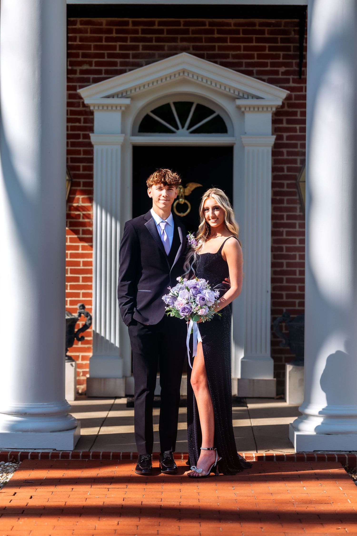 A couple stands together on a brick porch with white columns at a formal event wearing elegant attire.