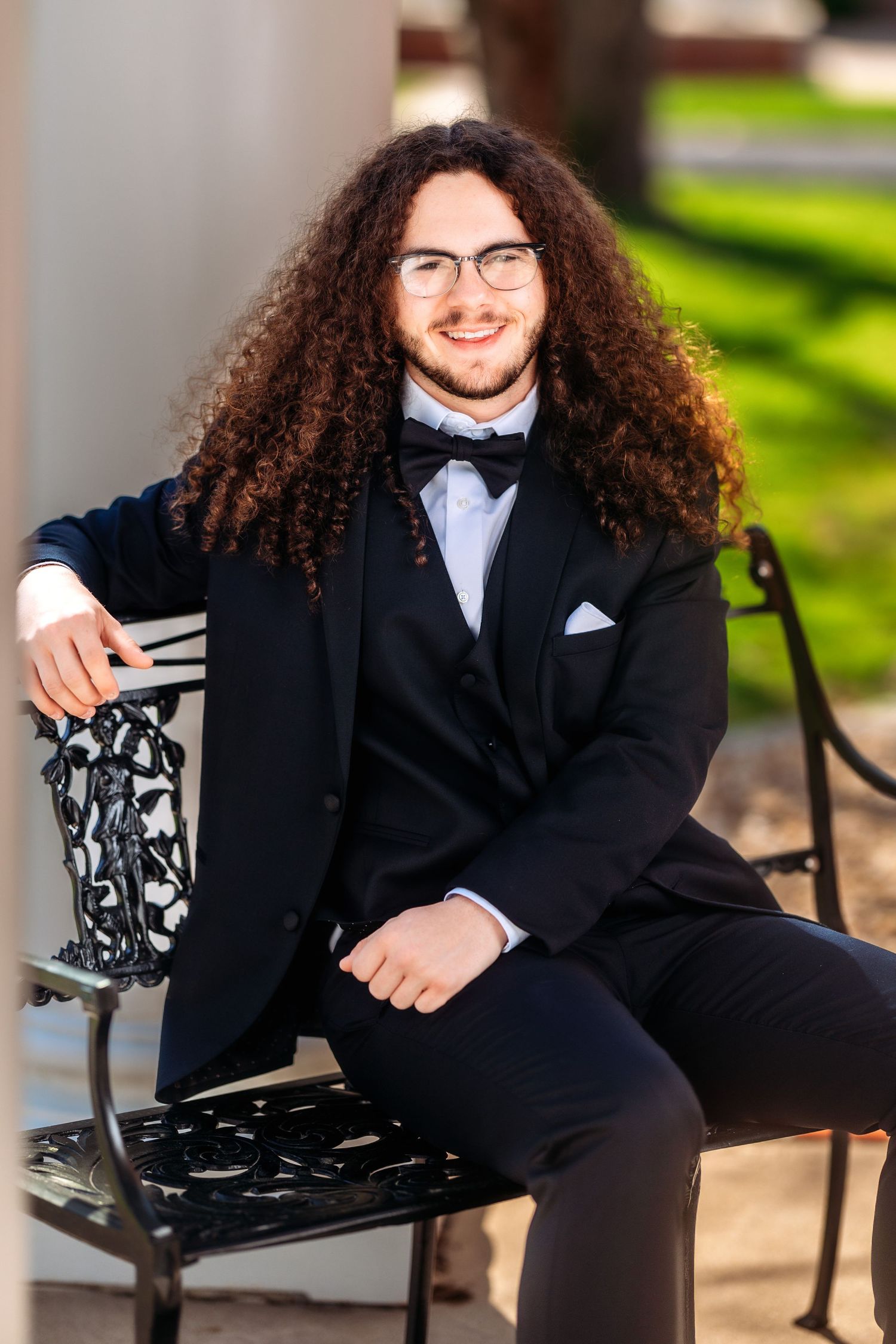 A person in a formal black suit and tie sits casually on an ornate metal bench in an outdoor garden setting.