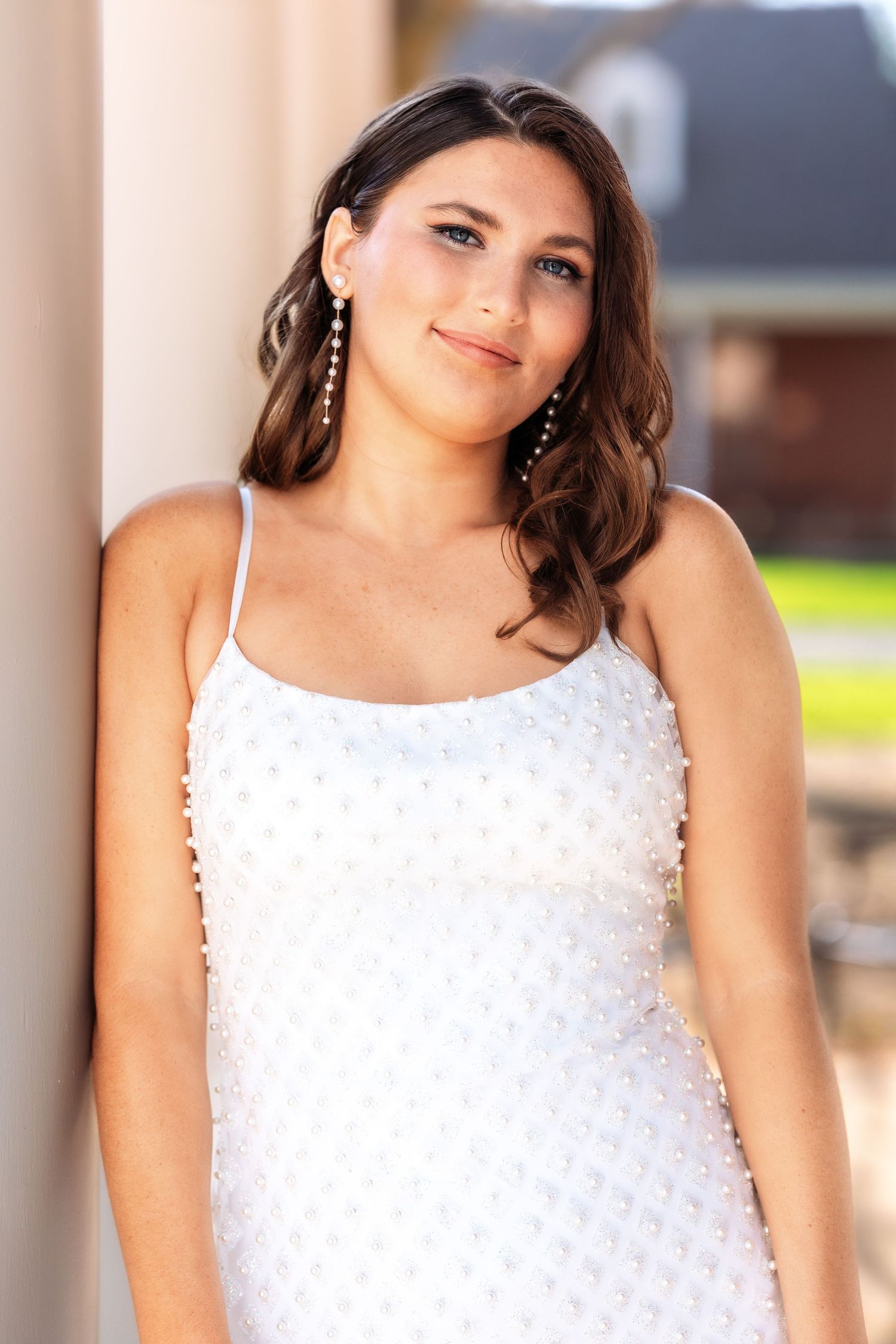 A woman in a white beaded spaghetti strap dress and dangling earrings poses against a light colored wall outdoors.