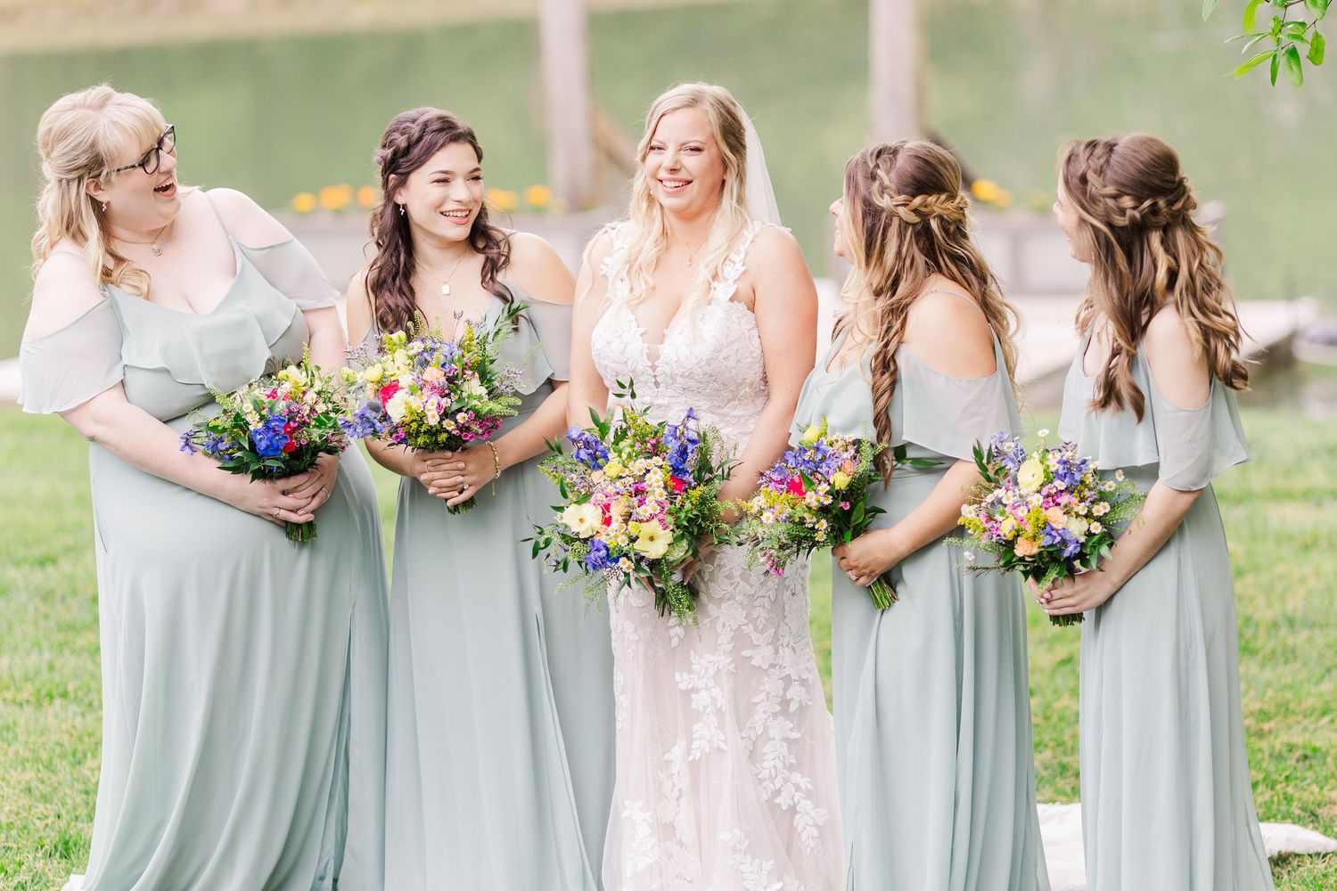 Wedding party in sage green dresses pose together holding colorful bouquets in a garden setting.