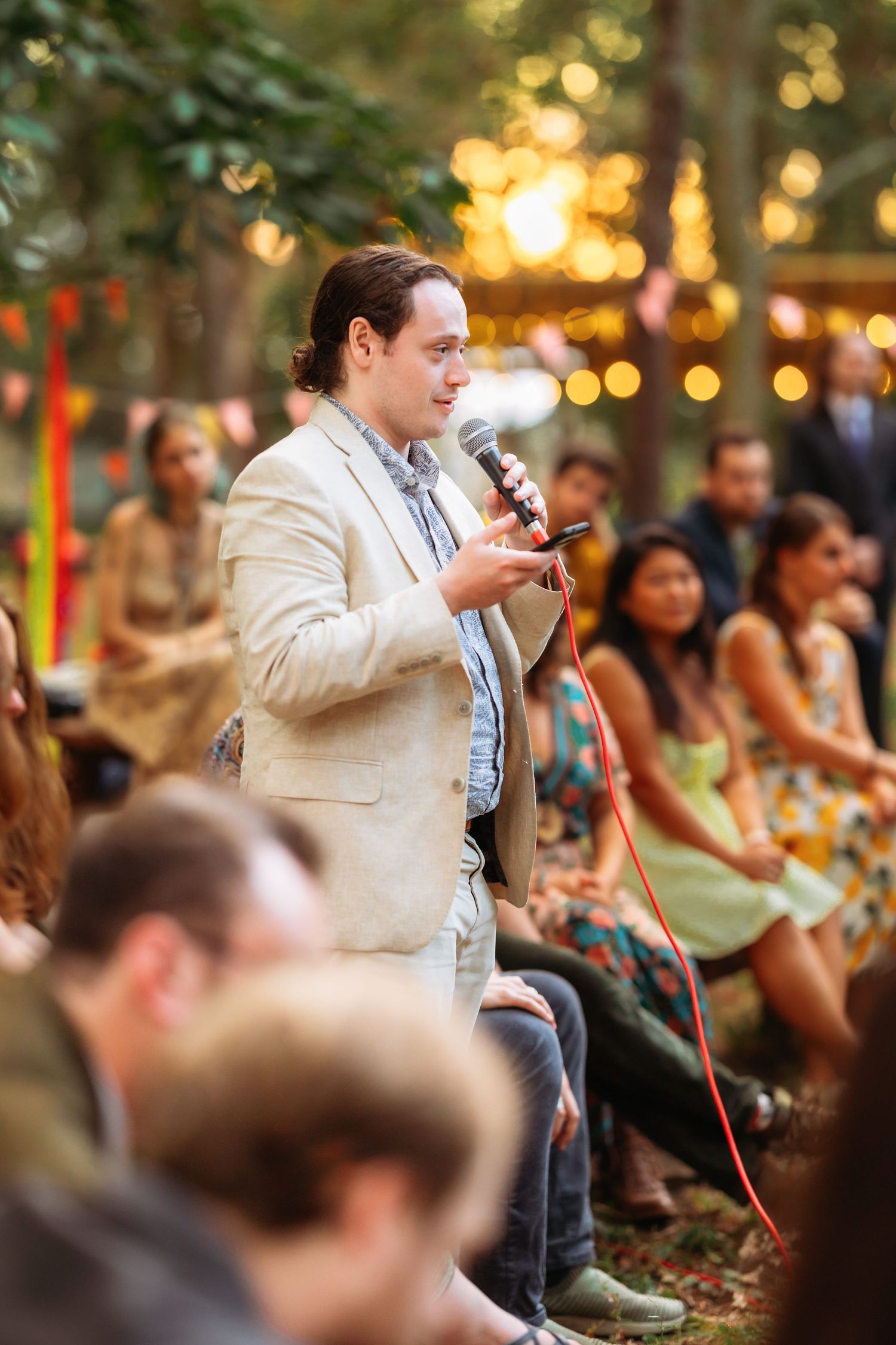 Speaker with microphone addresses wedding guests at outdoor reception with string lights.