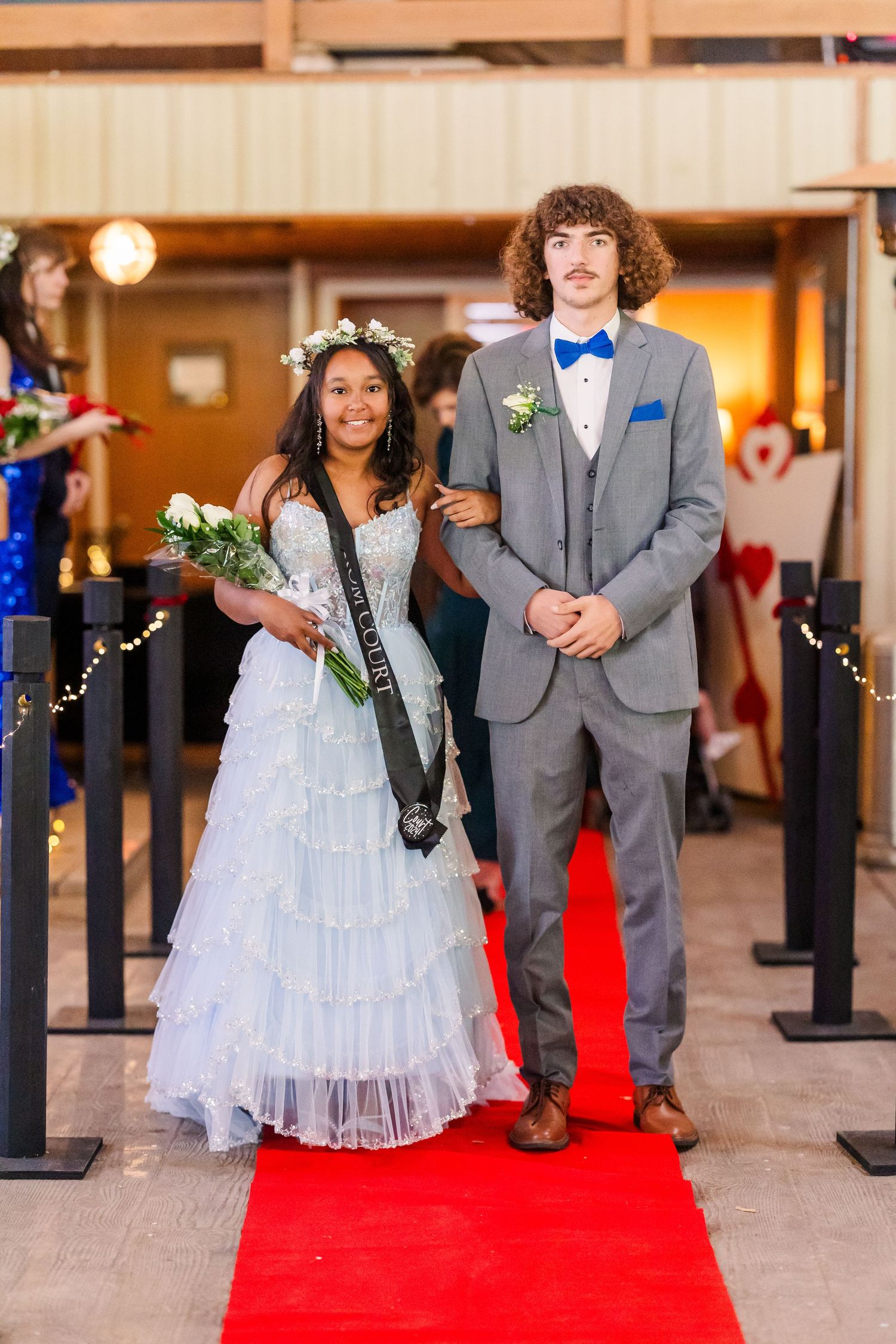 Couples walking down a red carpet in gray suits and light-colored dresses at a formal gathering.