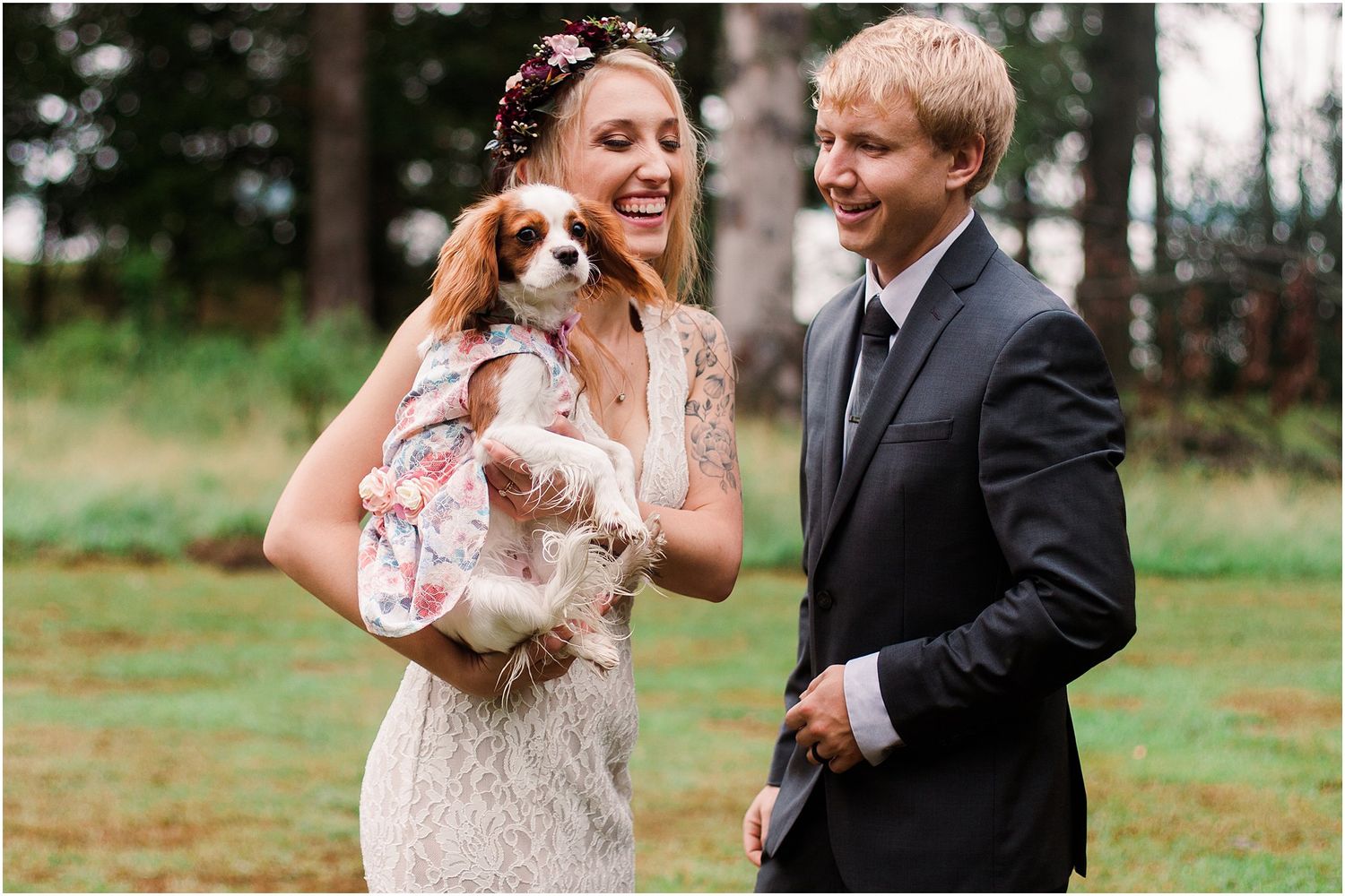 Wedding couple laughs while holding their small dog dressed in matching floral outfit.