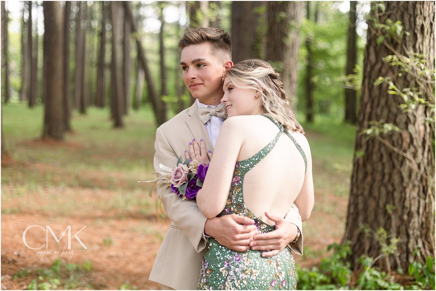A couple in formal attire embrace in a romantic forest setting with a green sequined dress and tan suit.