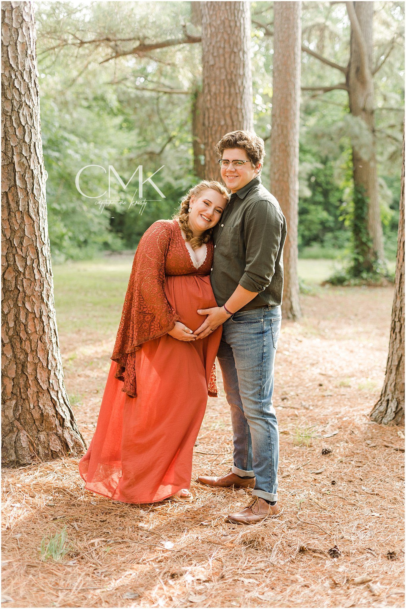 Expecting couple poses together among tall pine trees, wearing rustic autumn colors.