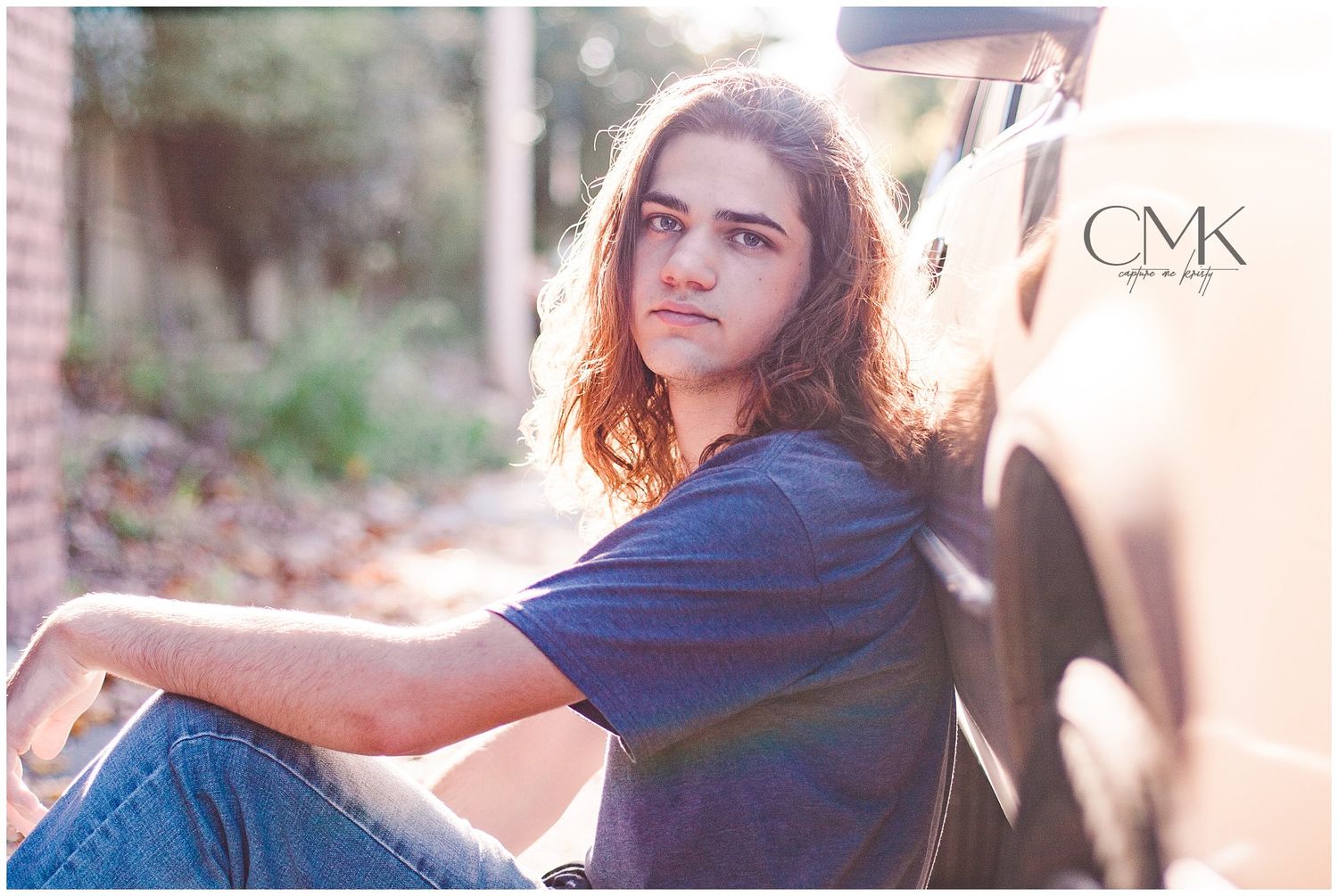 A relaxed outdoor portrait of someone sitting beside a car in casual attire.