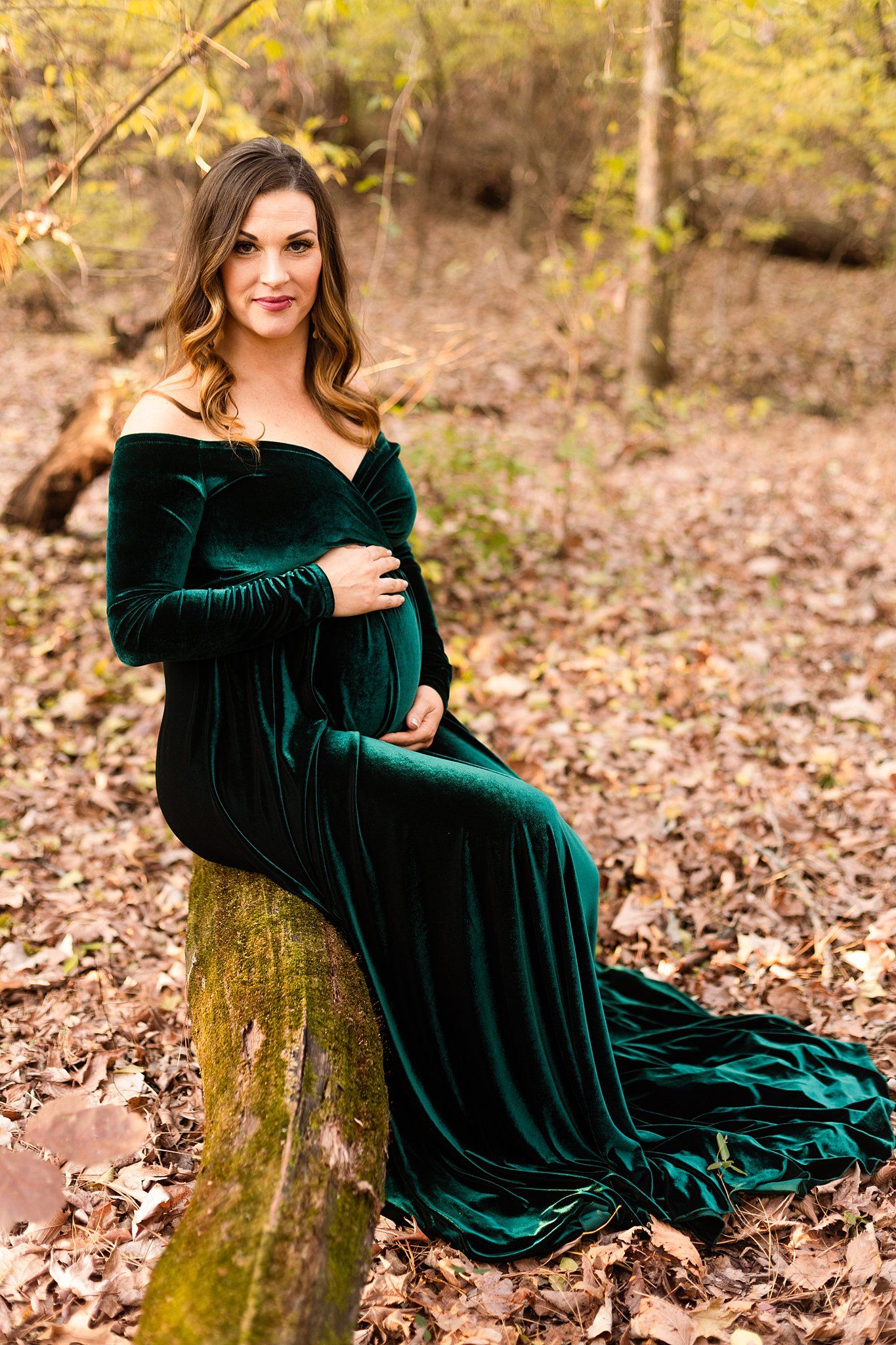 Maternity photo featuring emerald velvet gown on moss-covered log in autumn forest.