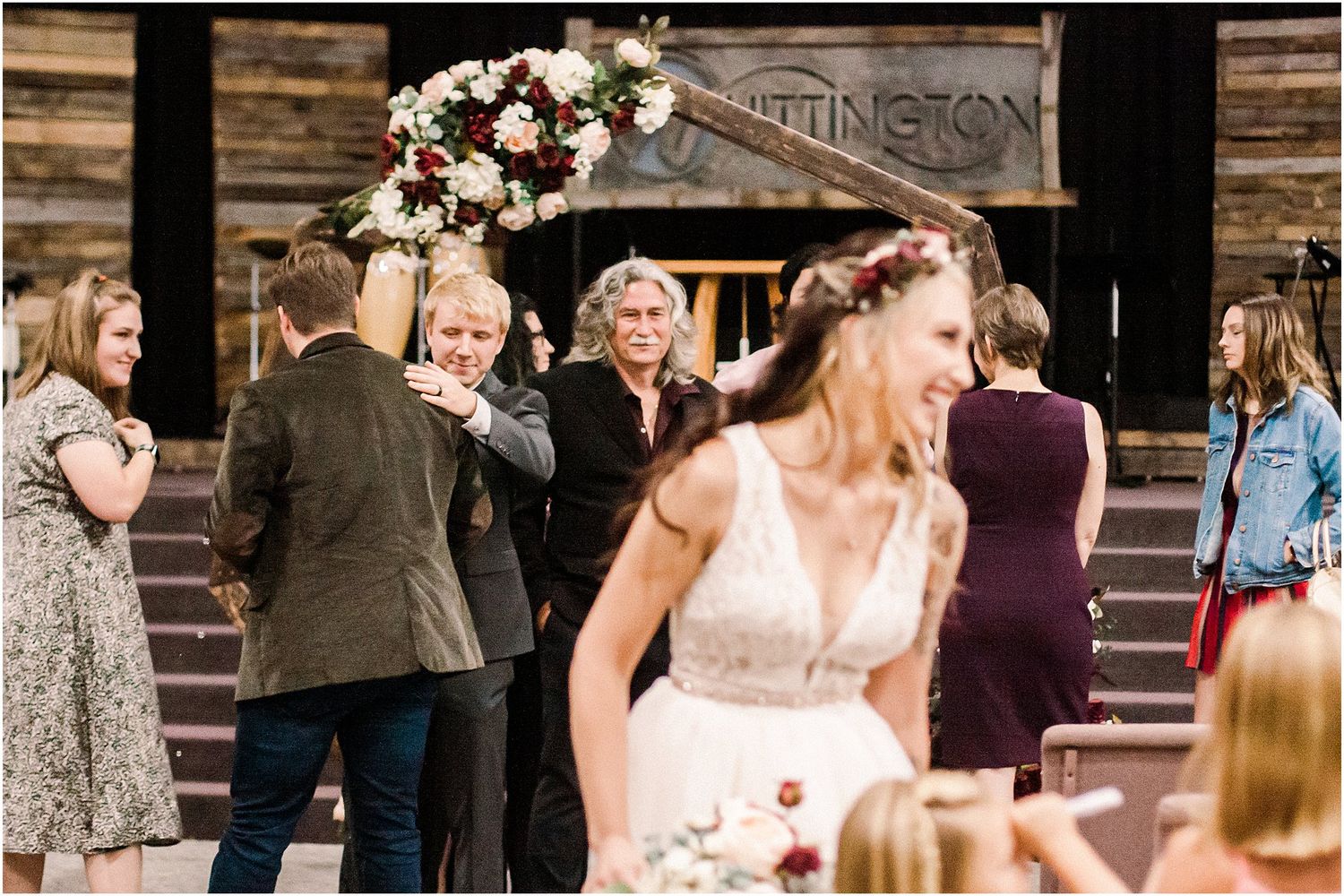 Wedding guests gather outdoors around a decorated entrance while celebrating at a rustic venue.