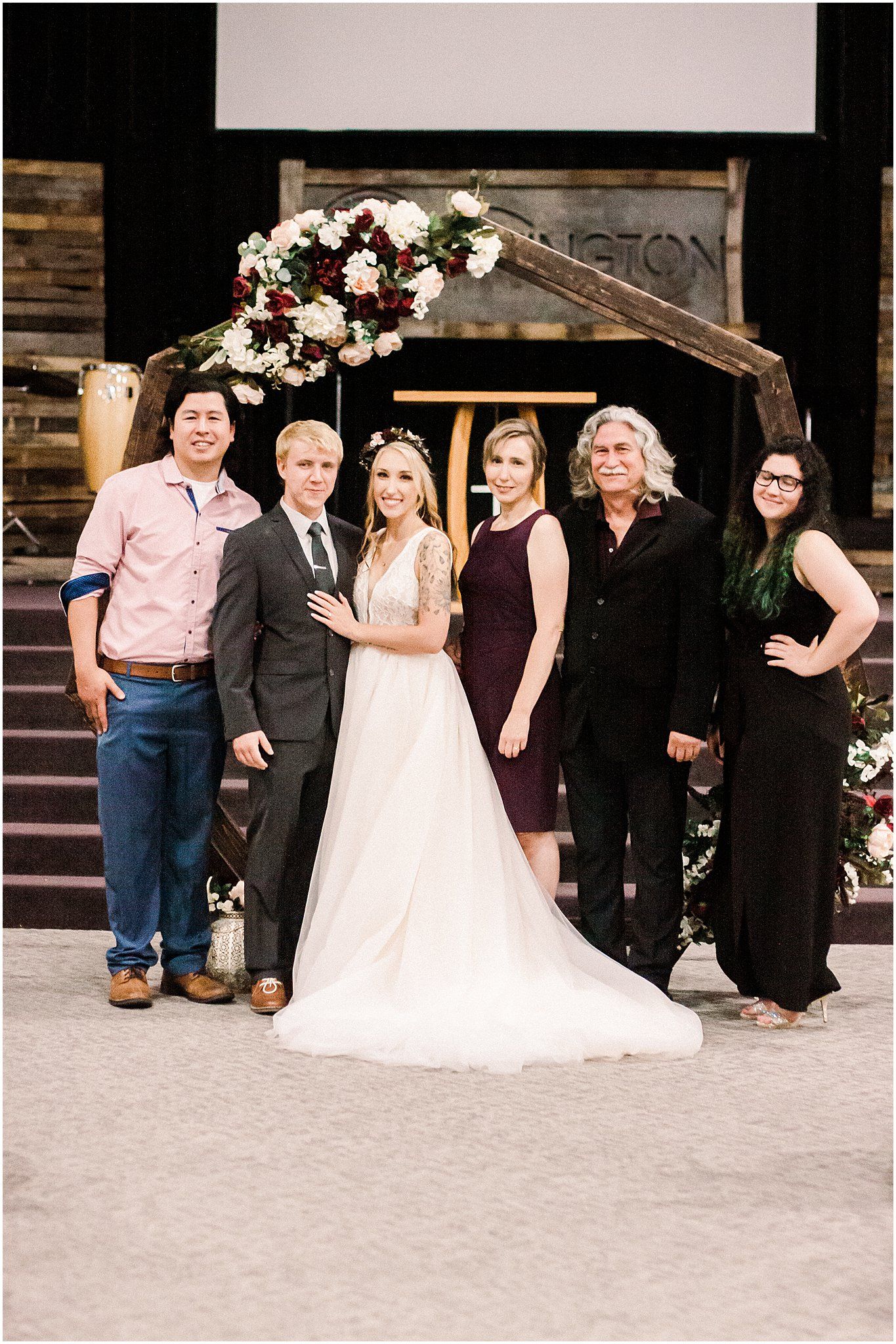 Wedding group photo under wooden arch with floral arrangements and rustic decor.