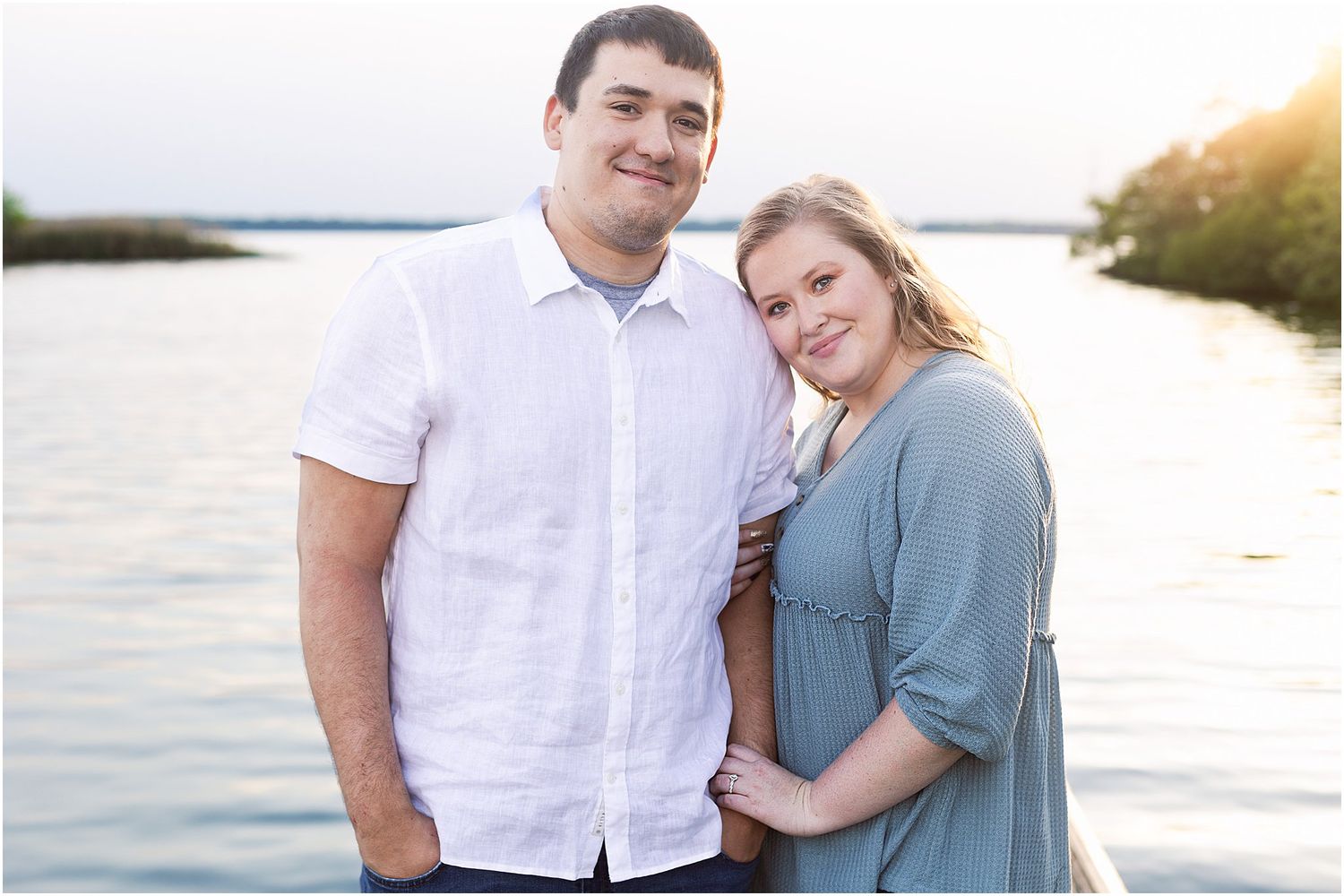 Couple poses together at waterfront during golden hour wearing white and blue casual outfits.