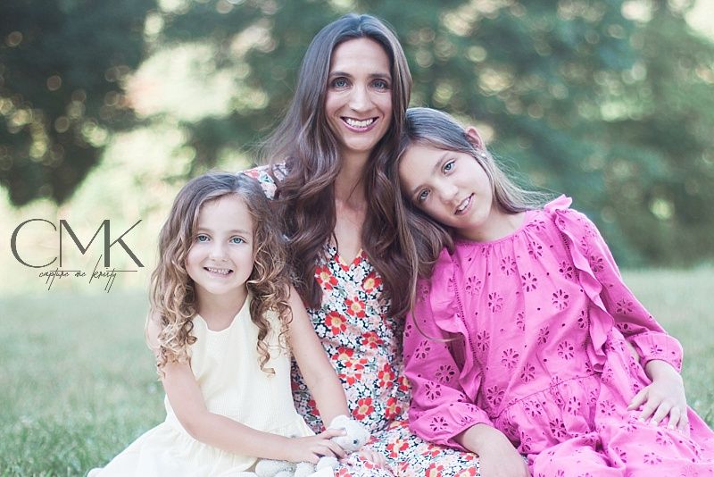 Family portrait of three in floral and pink dresses sitting on blanket outdoors with natural backdrop.