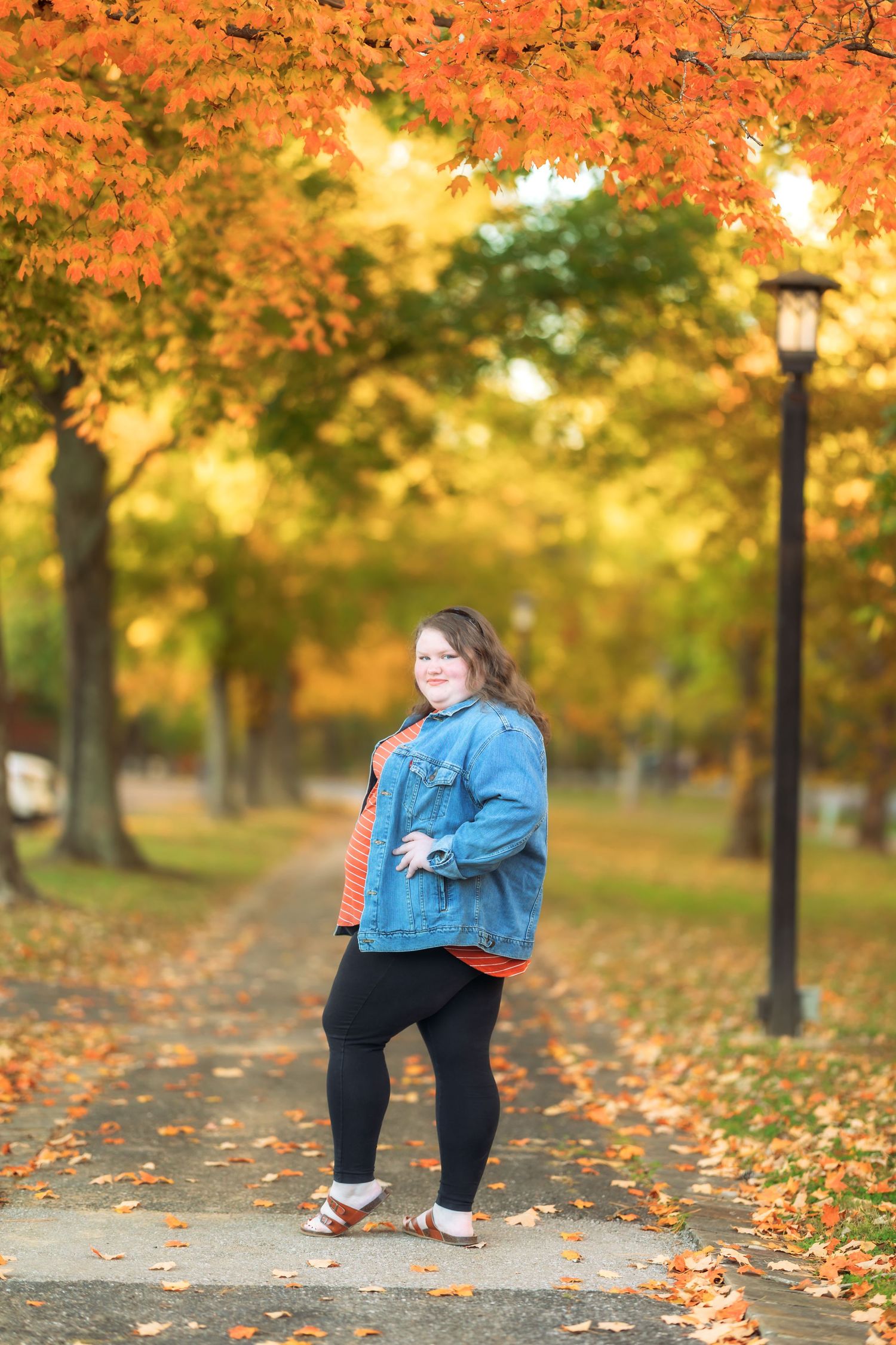 Person in denim jacket walks through park with golden autumn leaves.