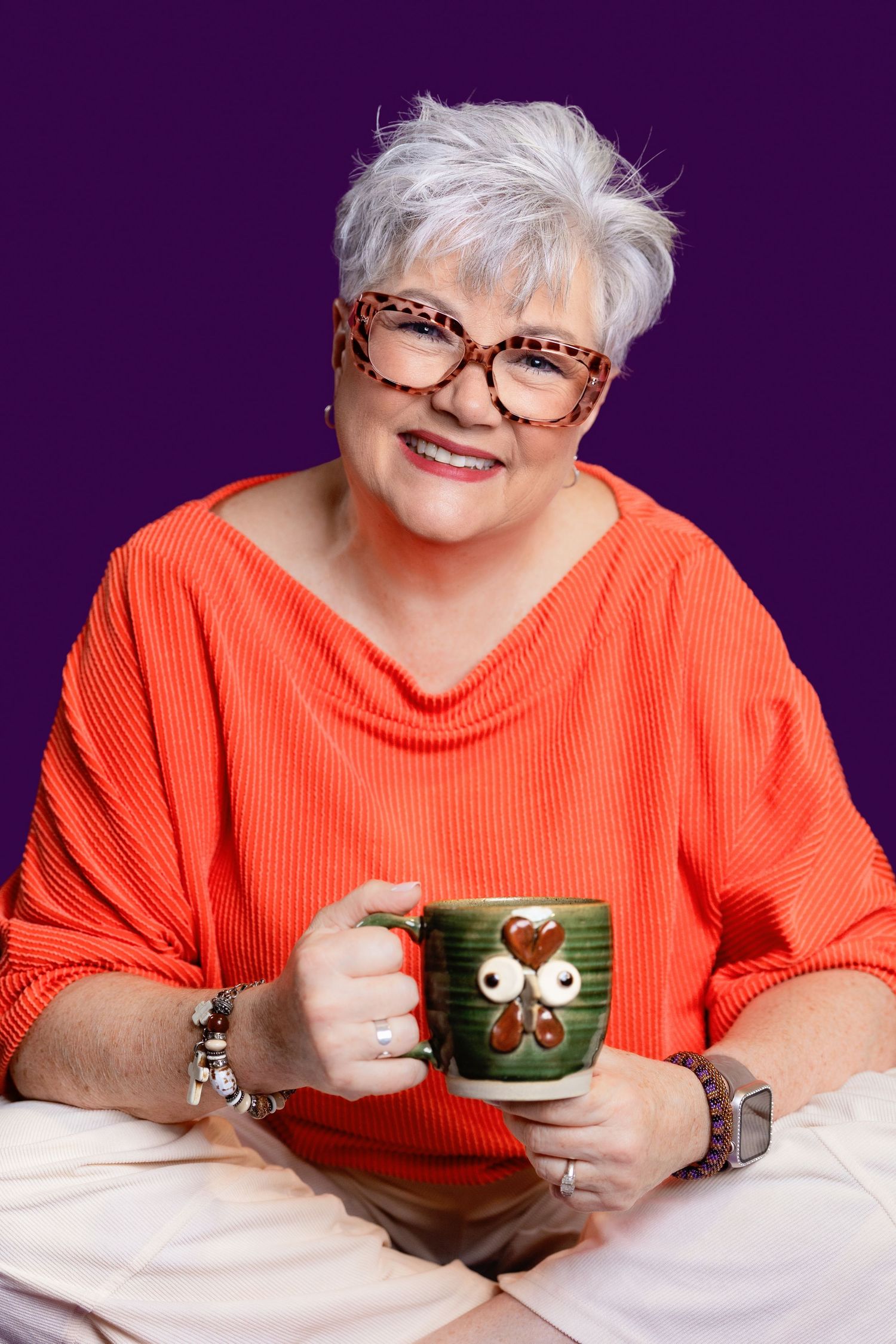 Smiling senior woman with short gray hair and leopard glasses holding a green mug, wearing an orange sweater.