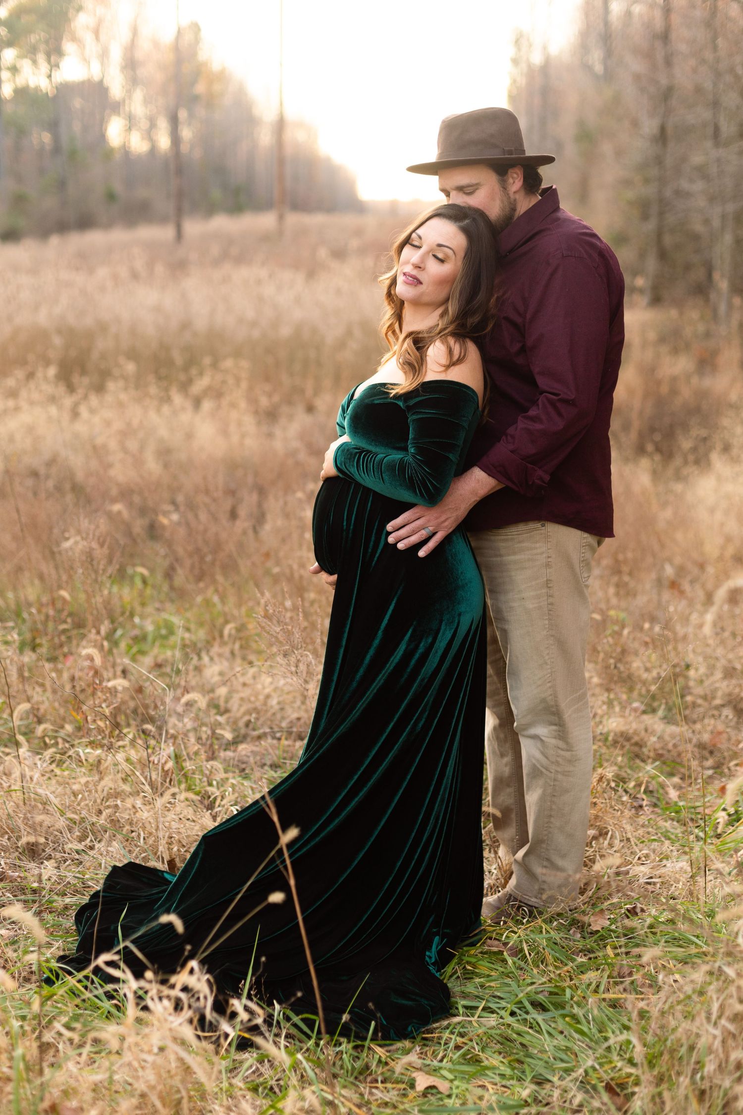 Couple embraces in field during maternity photo shoot with emerald velvet dress.