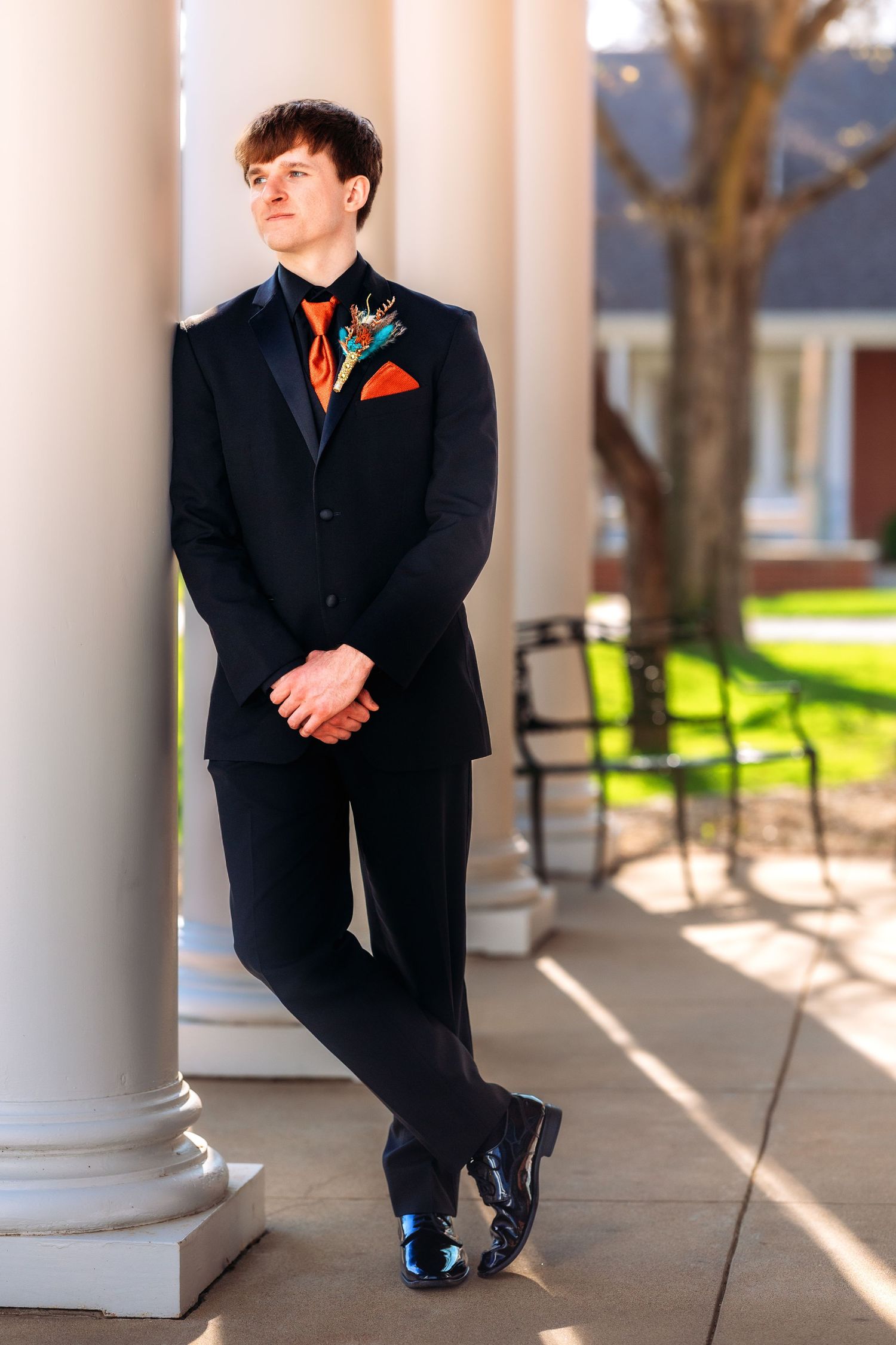 A person in a formal black suit with orange tie and pocket square leans against a white column on a sunny porch.