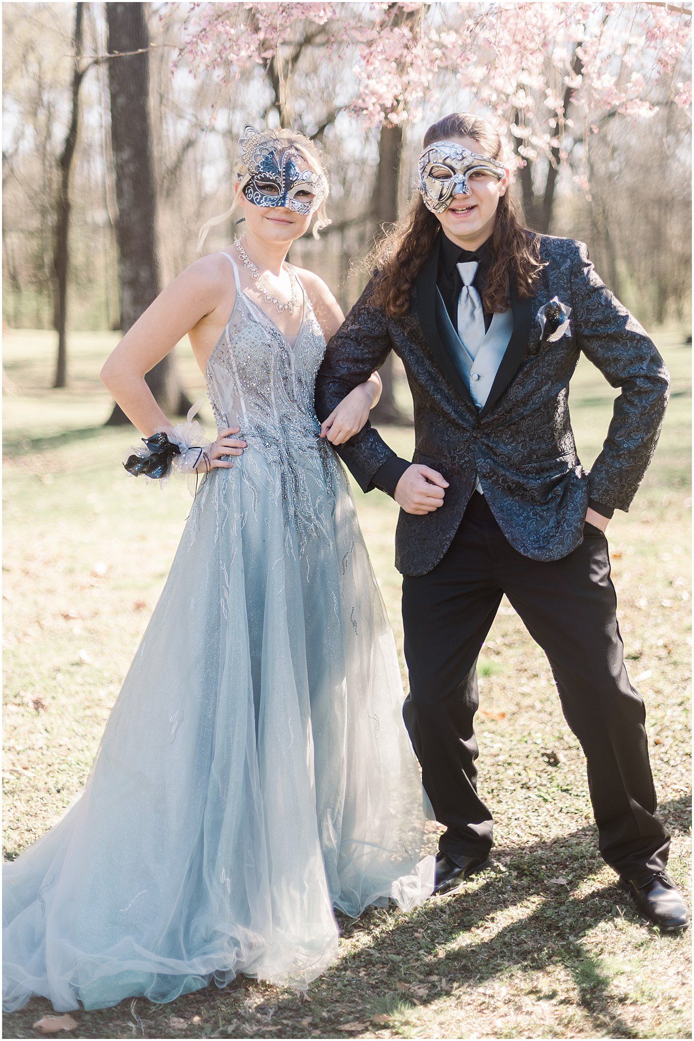 Formal couple in masquerade masks pose together under blooming cherry trees wearing blue gown and dark suit.