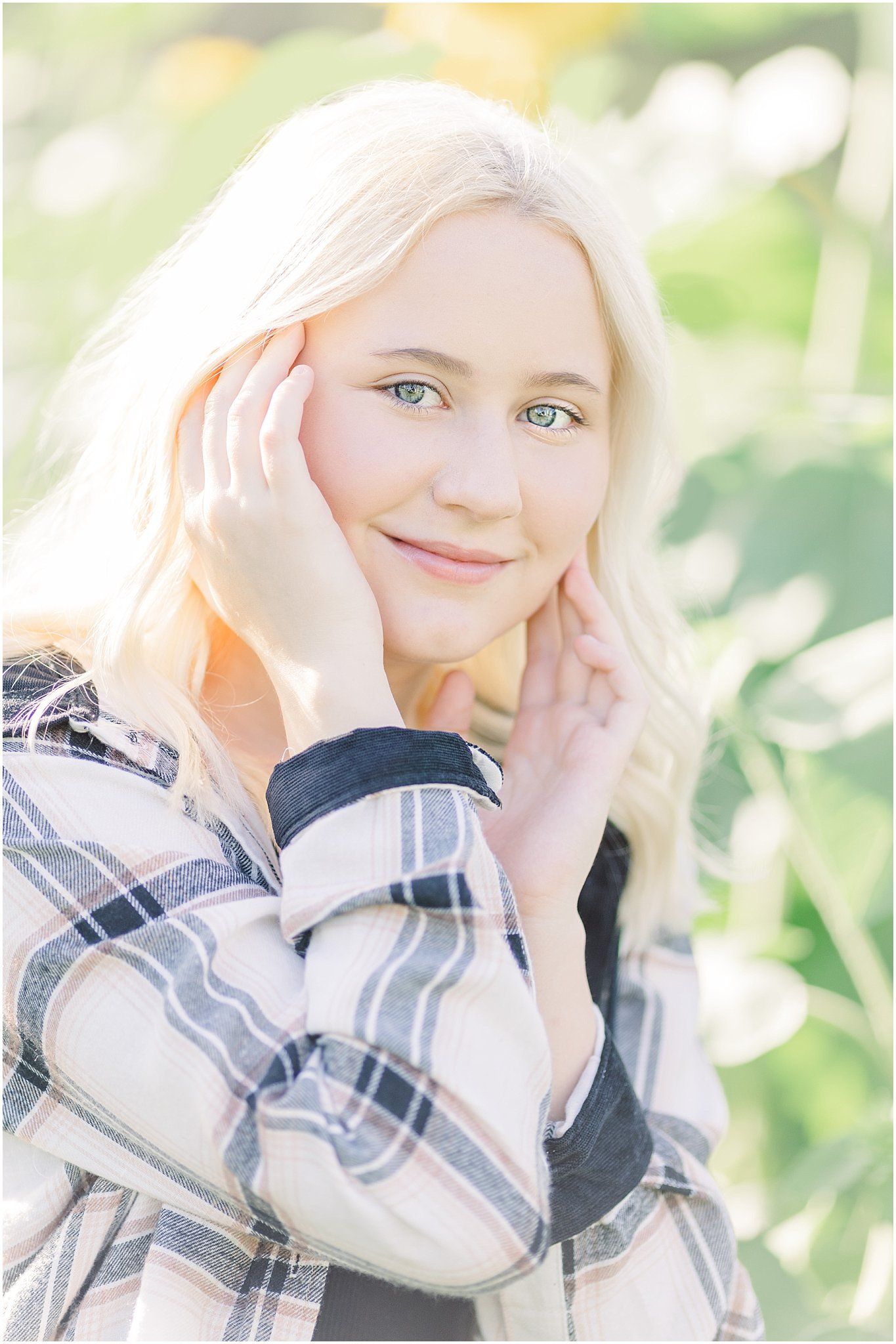 Senior girl delicately touches her face while kneeling in sunflower field