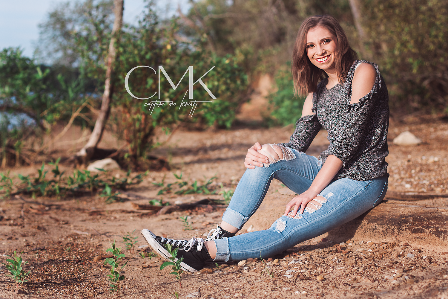 Casual outdoor portrait of woman in grey top and jeans sitting on rocky ground.